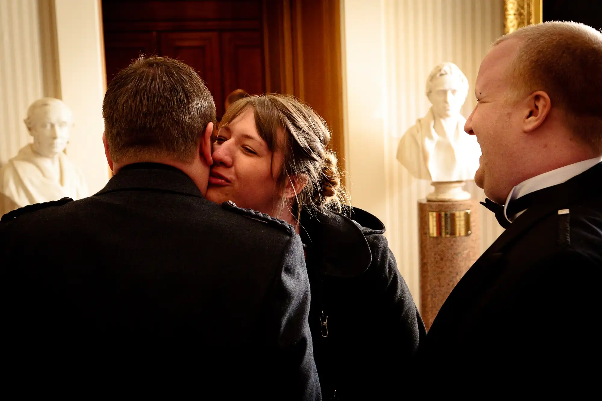 A woman kisses a man on the cheek inside a warmly lit room adorned with bust sculptures, while another man looks on, smiling.