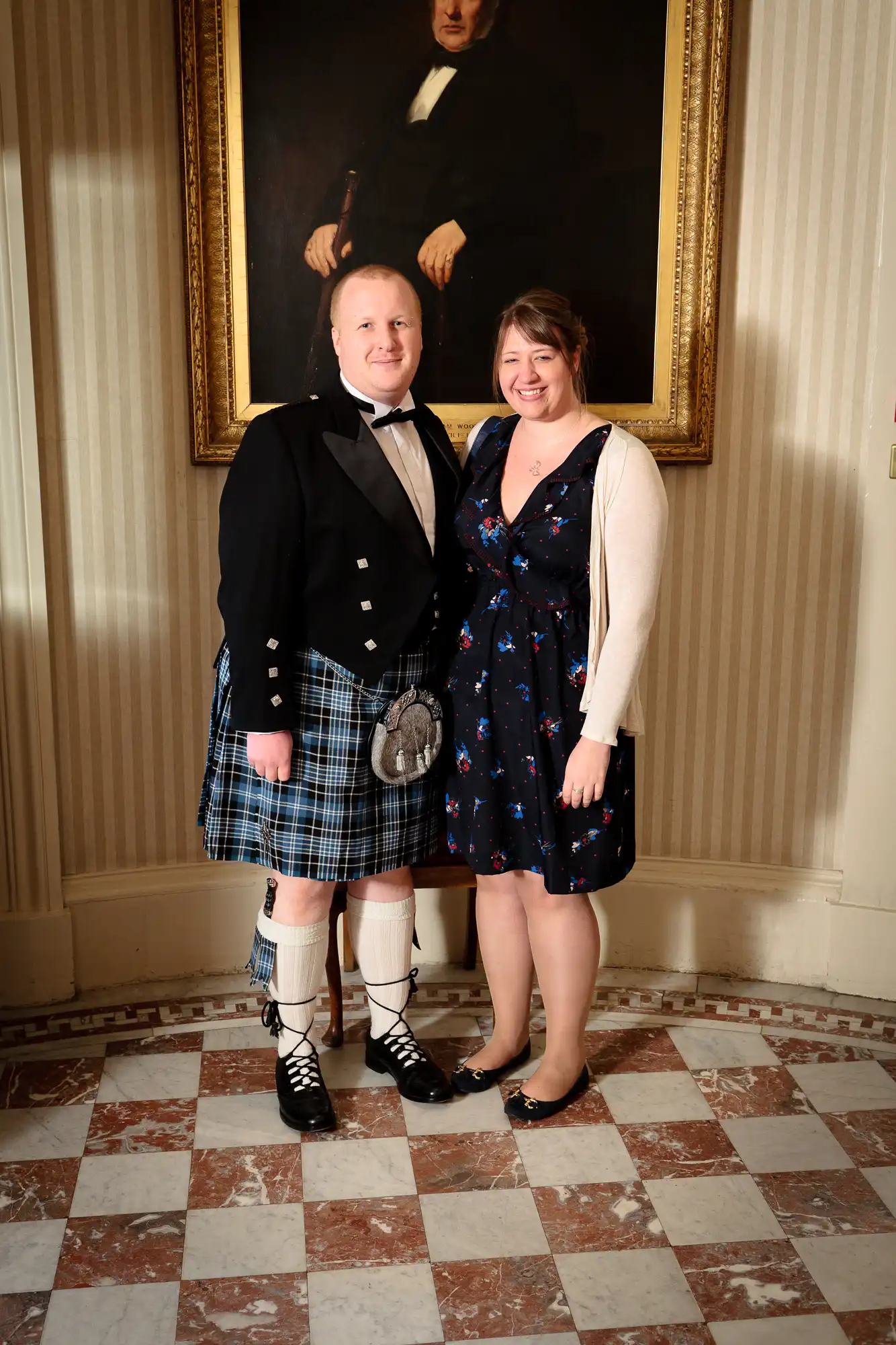 A man in a traditional kilt and a woman in a blue floral dress smiling while standing in front of a portrait in an elegant room.