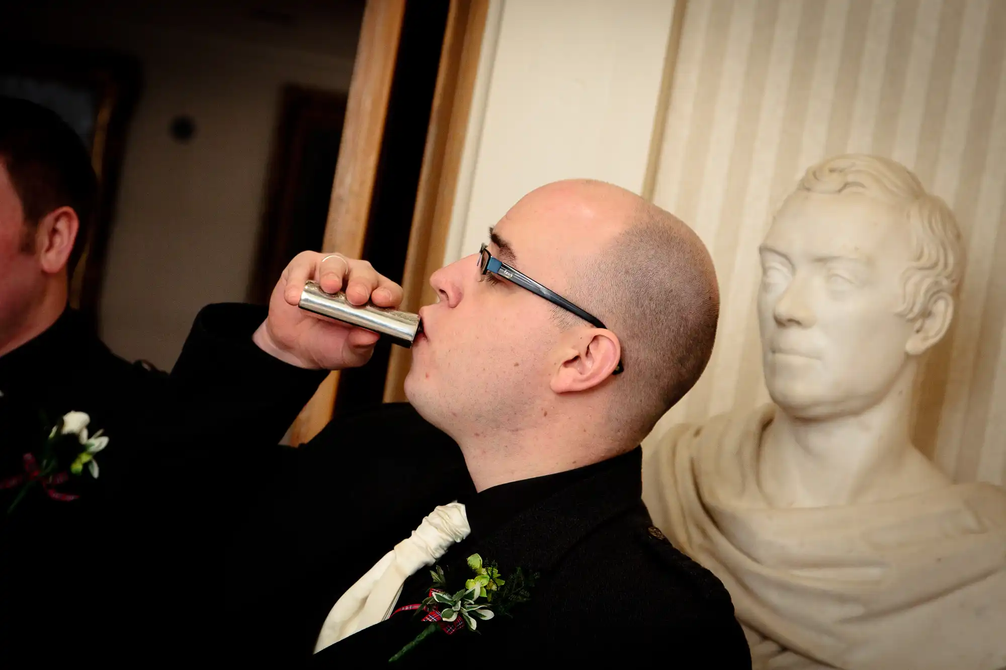 A man in a tuxedo with a boutonniere drinks from a flask, a bust sculpture in the background.