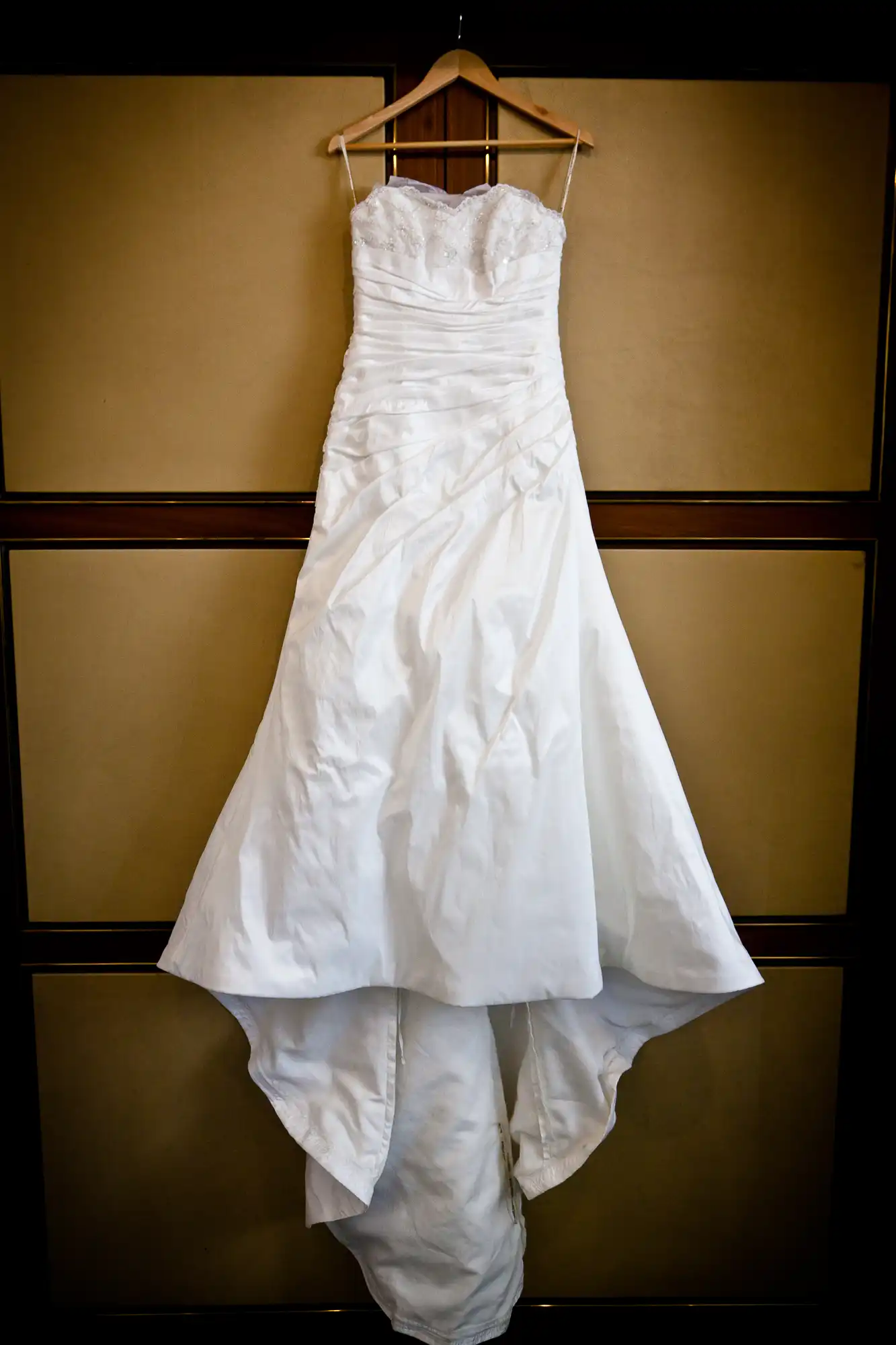 An elegant white wedding dress with a sweetheart neckline hangs on a wooden hanger against a patterned beige background.