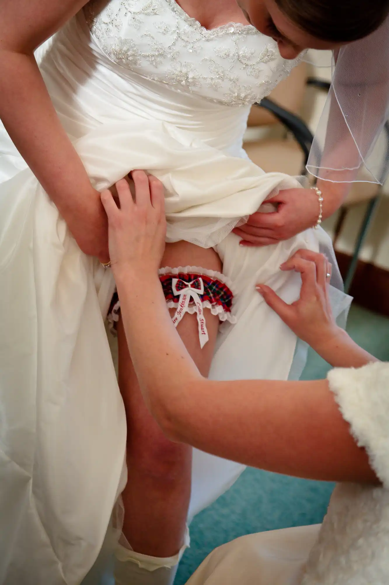 A bride adjusts a red and white garter on her thigh while another person assists, focusing on the details of her elegant white wedding dress.