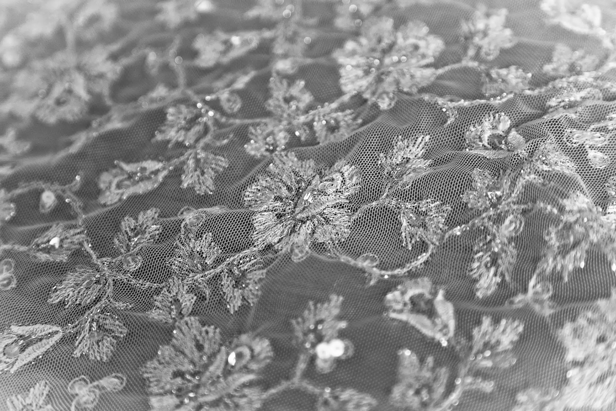 Close-up of a delicate lace fabric with intricate floral embroidery and beadwork in grayscale.