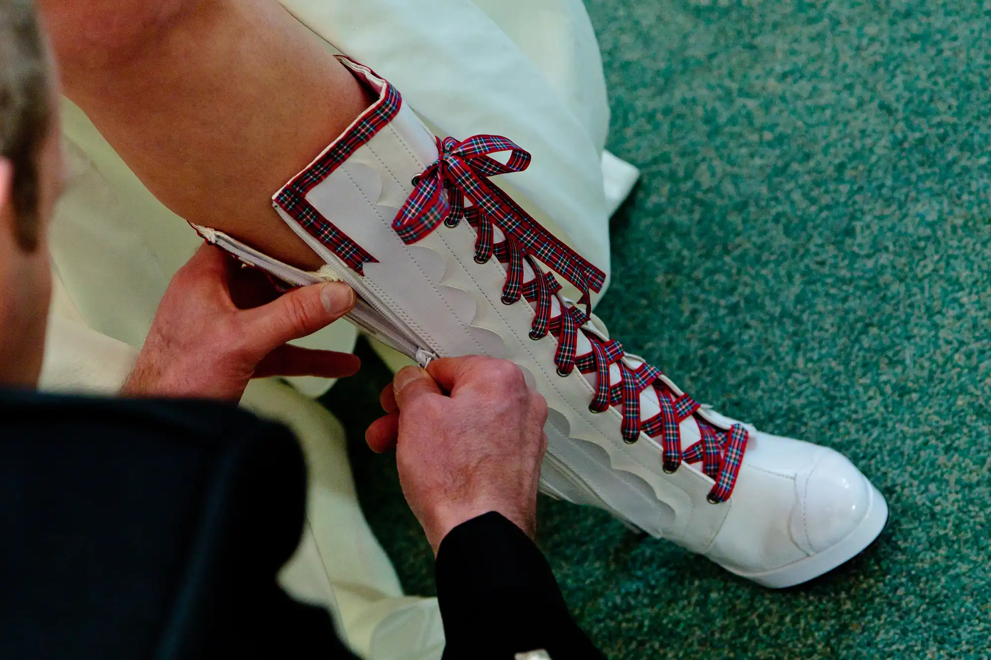 A person ties the red and blue plaid laces on high-top white boots.