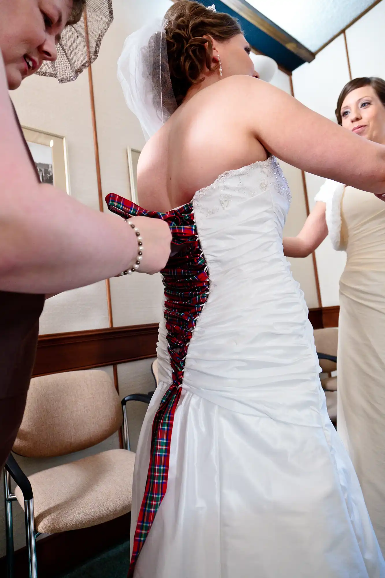 A bride being assisted with her dress by two women, featuring a white gown with red and plaid corset lacing in a room with beige chairs.