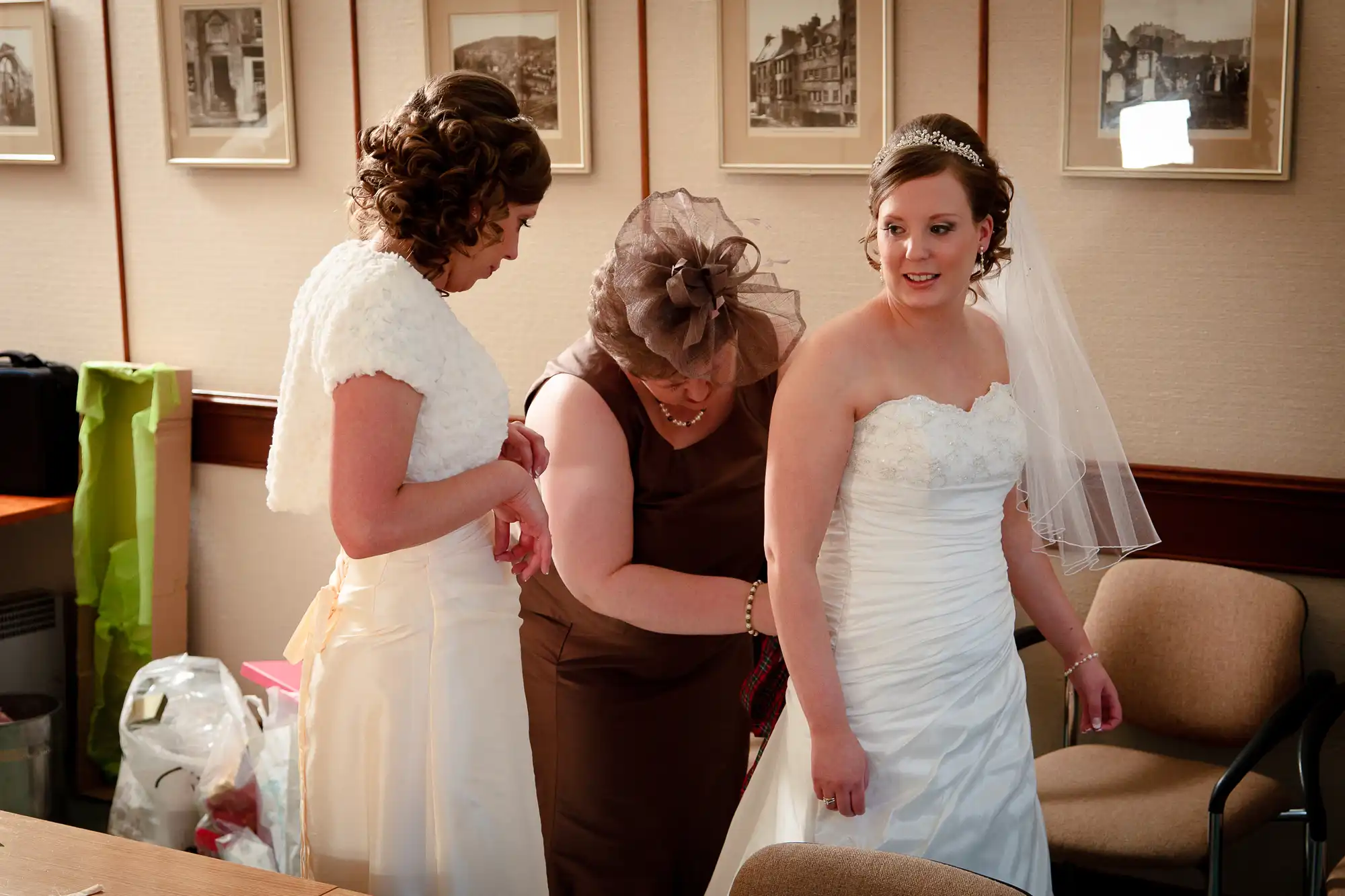 Two women adjusting a bride's wedding dress in a room, with emotional expressions on their faces.