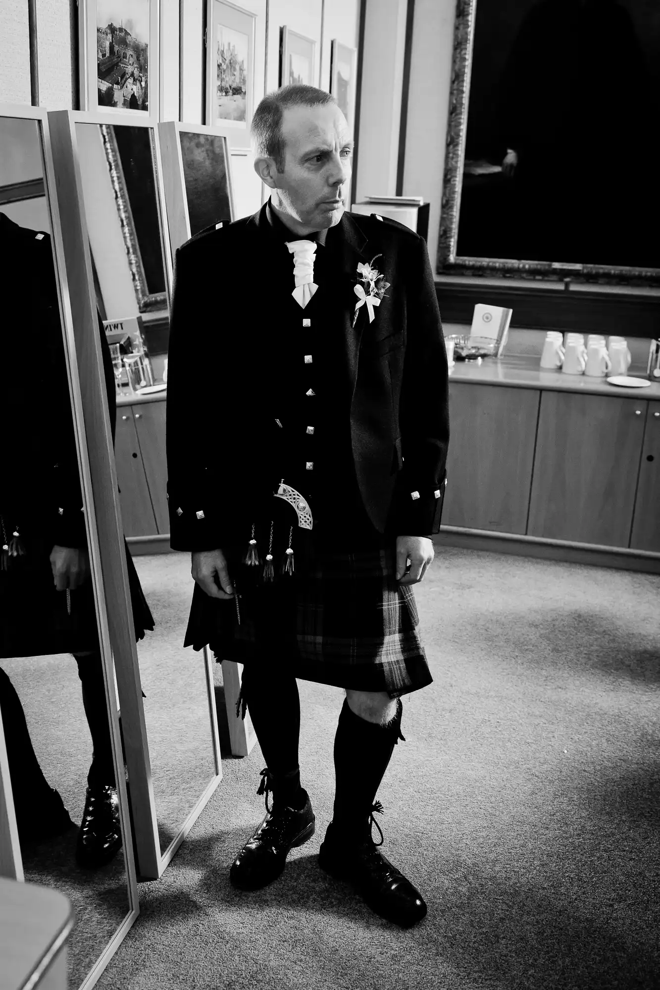 A man in a formal kilt outfit with a sash and medals stands pensively in front of a mirror in a black and white photo.