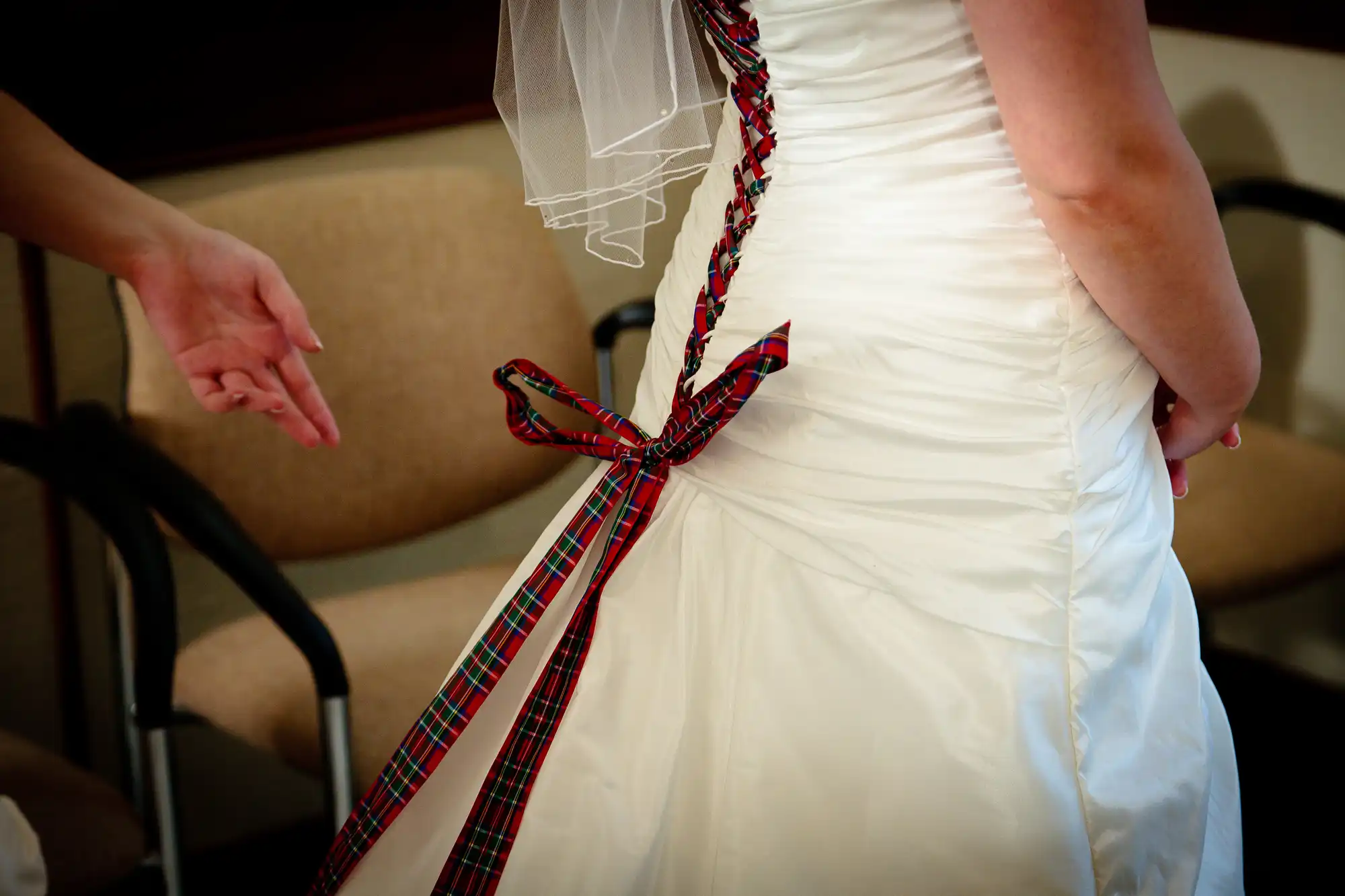 A bride in a white dress with a tartan ribbon tied at her waist extends her hand towards someone off-camera.
