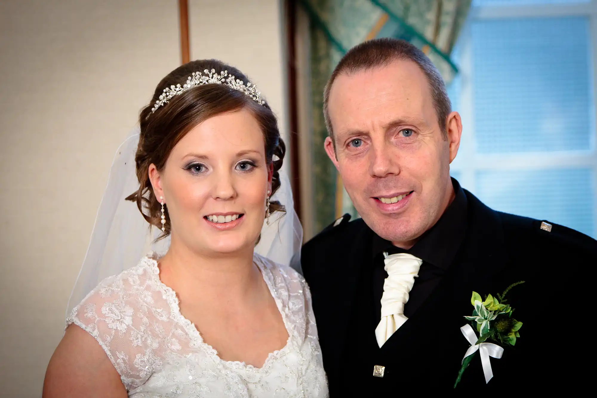 A bride and groom smiling in formal wedding attire, with the bride wearing a lace dress and tiara, and the groom in a black suit with a white tie and boutonniere.