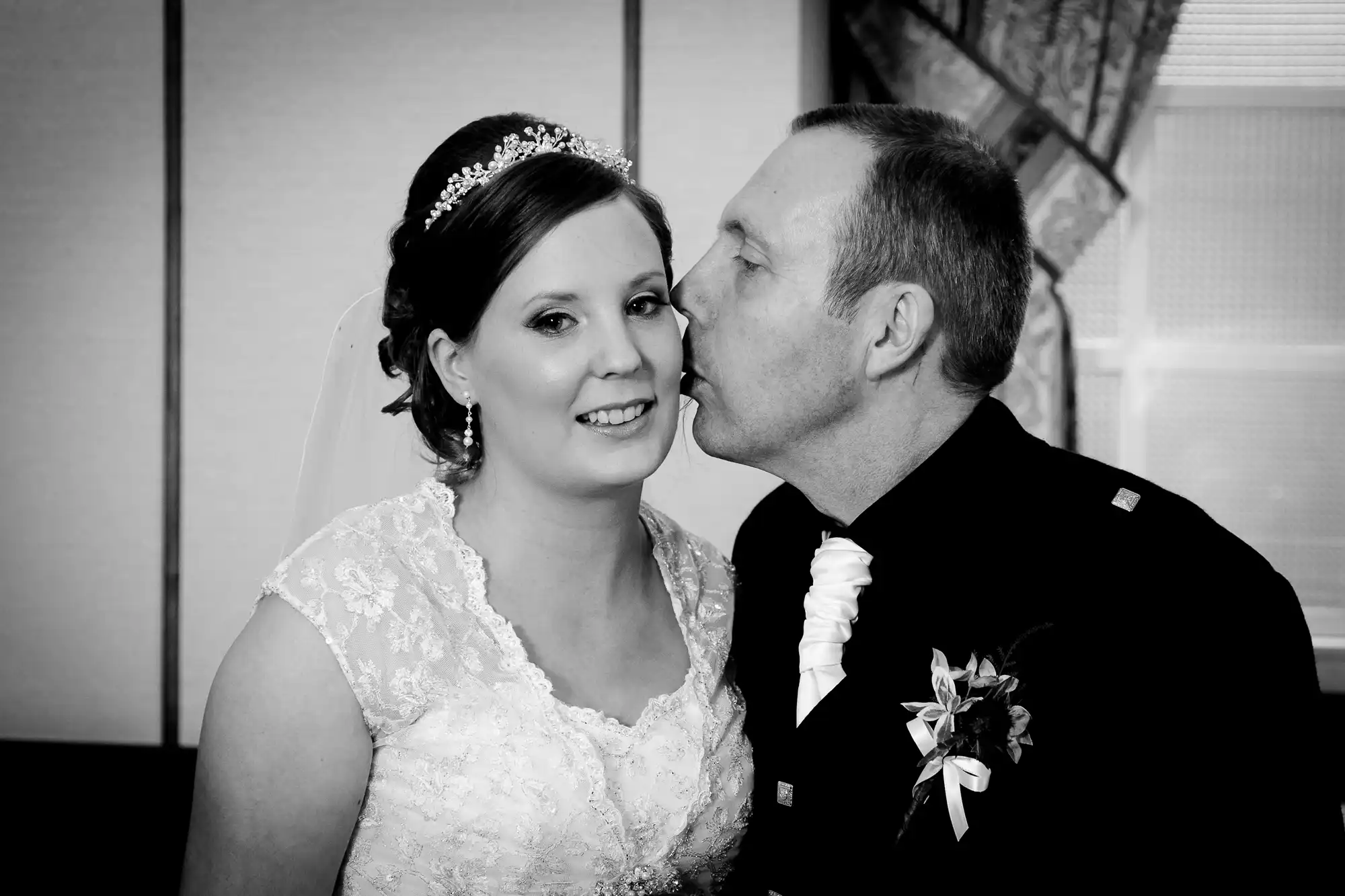 A black and white photo of a bride and groom kissing, with the groom in a tuxedo and the bride wearing a lace dress and tiara.