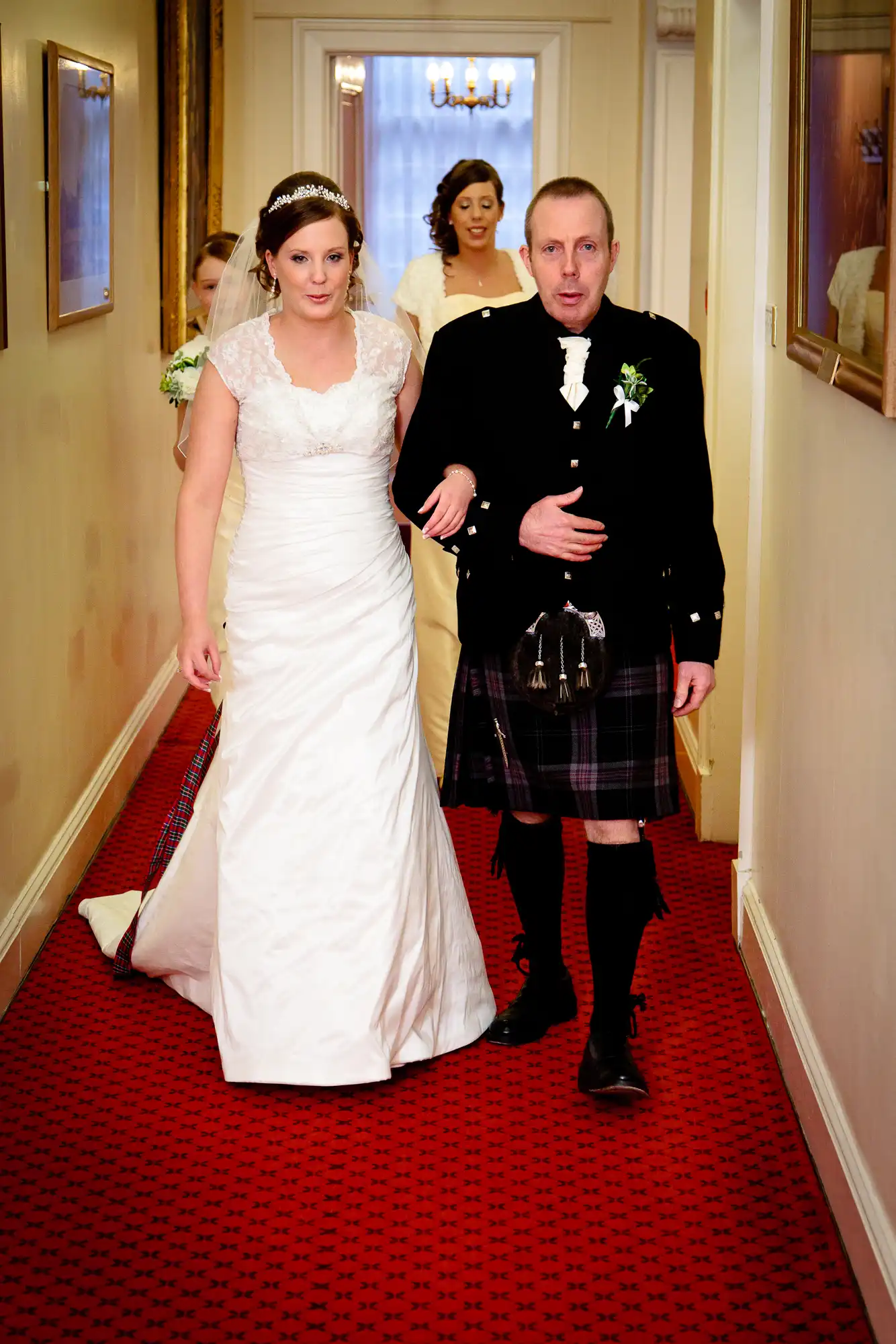 A bride in a white dress and a groom in a kilt and jacket walk down a hallway with red carpet, followed by a bridesmaid in a blue dress.