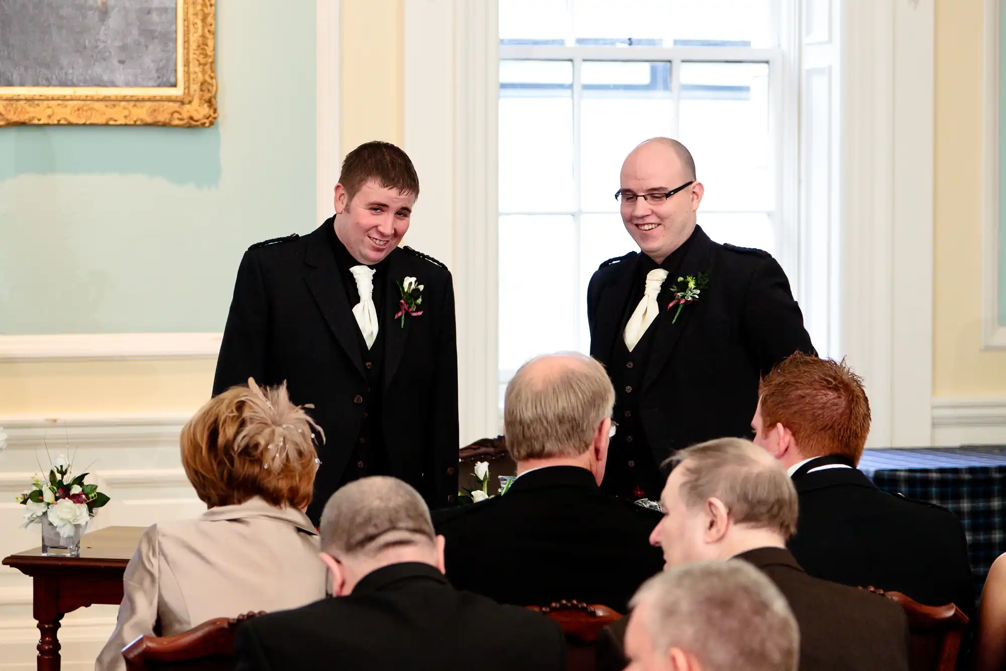 Two men in tuxedos, smiling during a wedding ceremony in a room with guests and elegant decor.