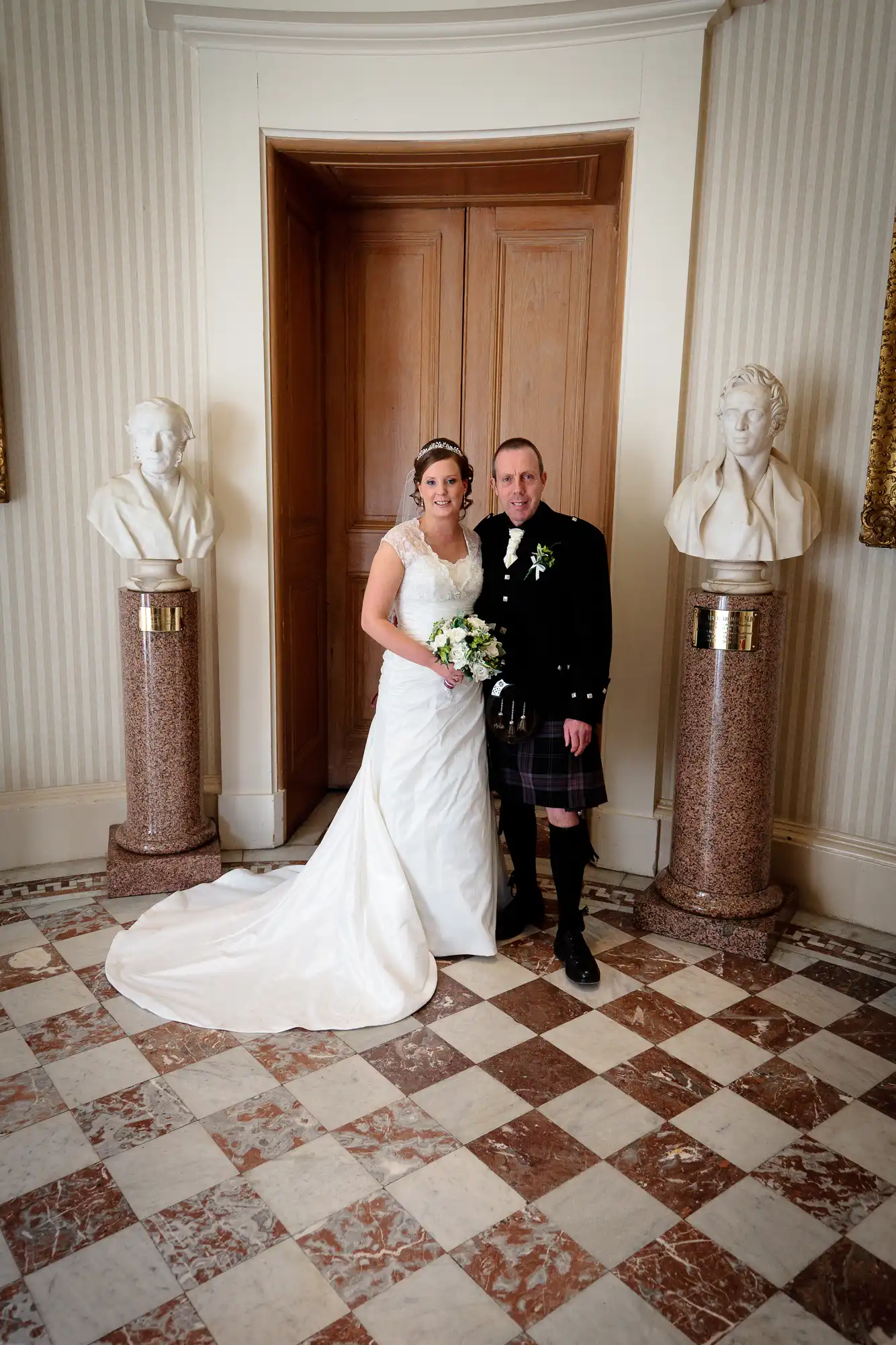 A bride in a white dress and a groom in a kilt posing together between two bust sculptures in an elegant room with striped wallpaper and a tiled floor.
