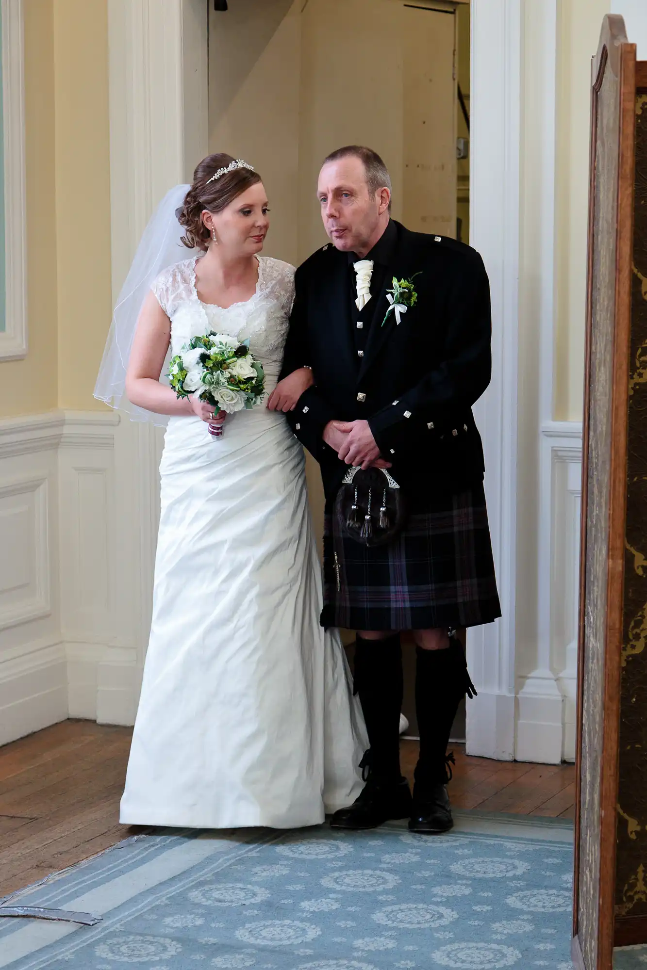 A bride in a white dress and a groom in a kilt standing together in a doorway, both looking away from the camera.