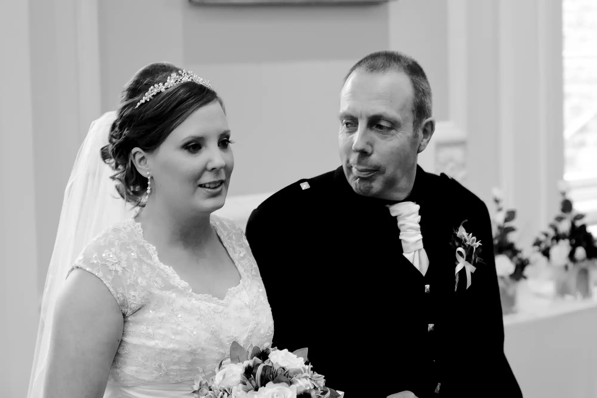 A bride in a white lace gown and a man in a black suit with an orange ribbon pose together, smiling in a black and white photo.