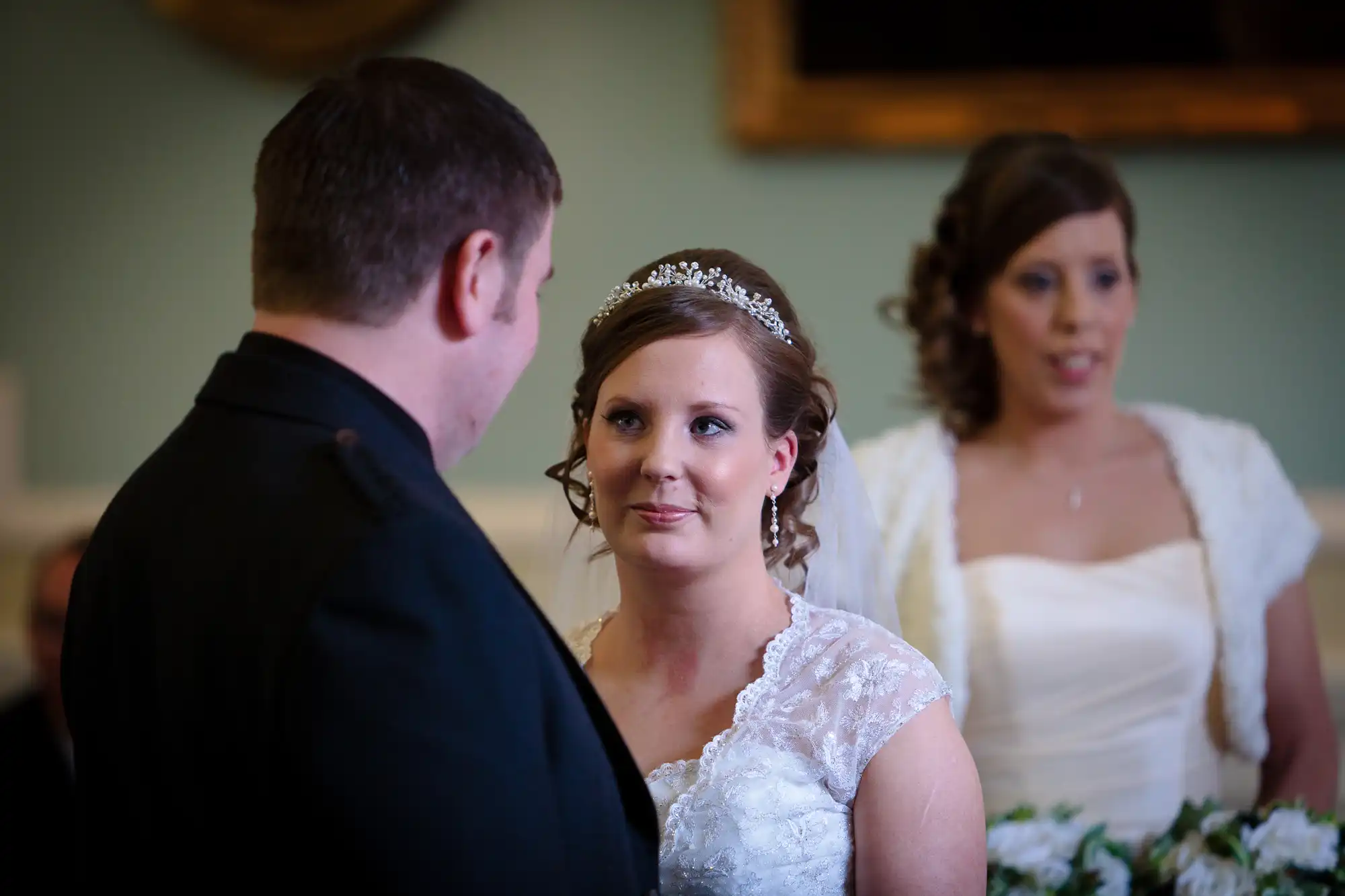 Bride in a lace dress and tiara gazes at groom with a focused expression, another woman in bridal attire in the background.