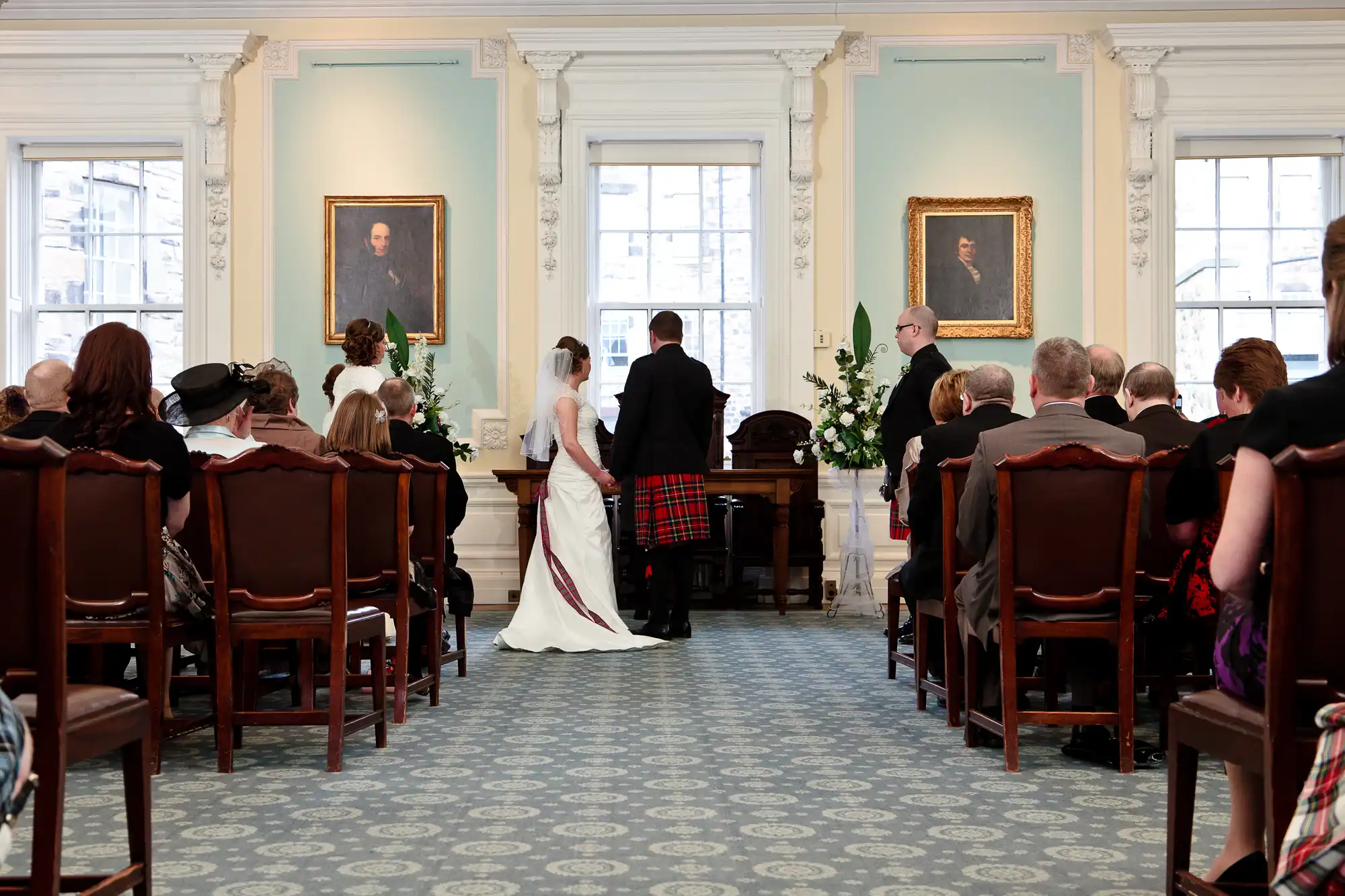 A wedding ceremony in an elegant room with guests seated, the bride and groom standing at the altar, and portraits on the walls.