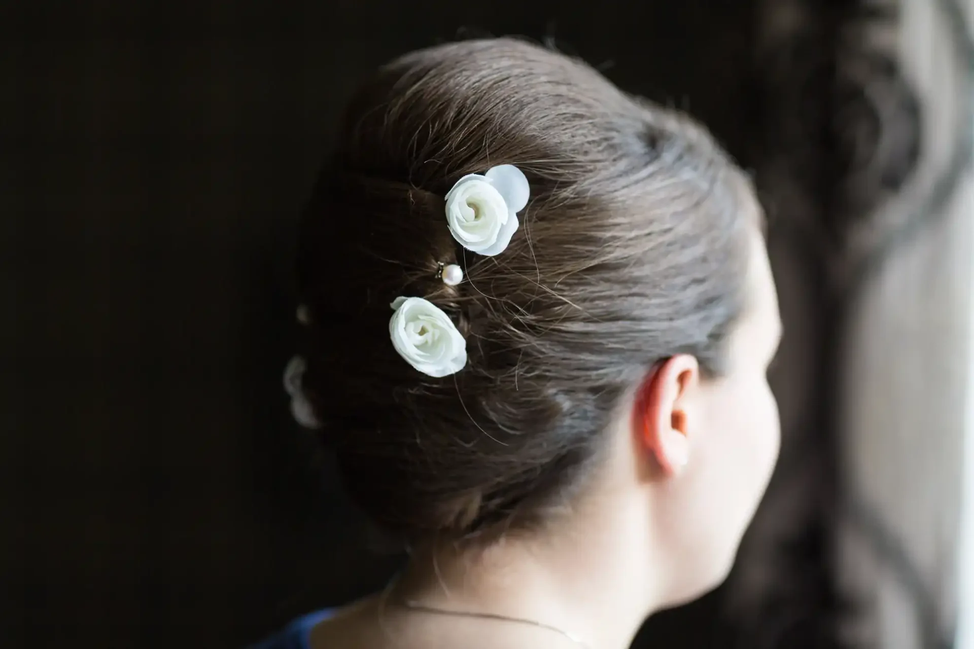 Rear view of a woman's hairstyle with three white rose accessories and tiny pearls, elegantly styled in a bun.