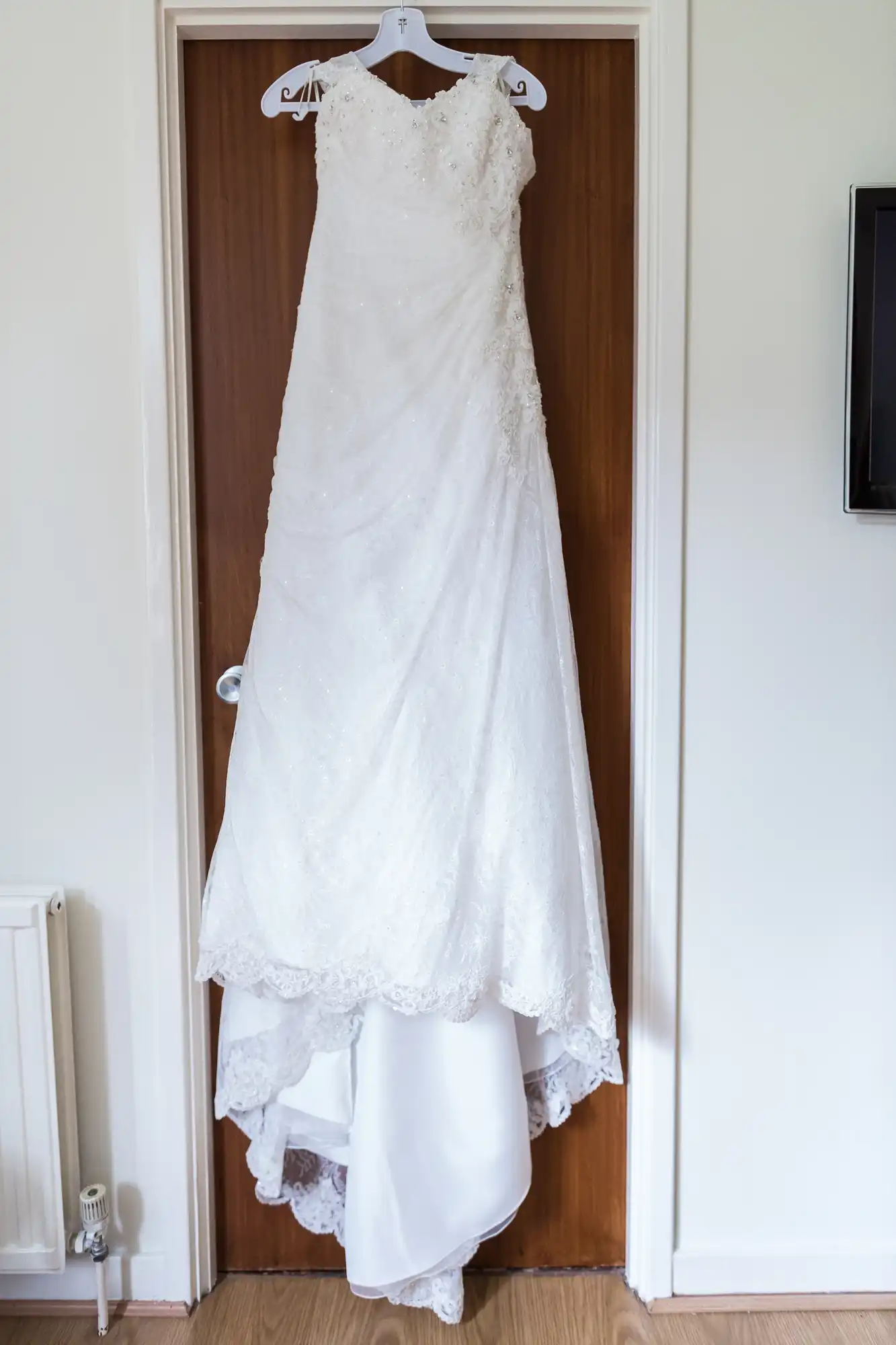 A white wedding dress with lace detailing hanging on a wooden door in a well-lit room.