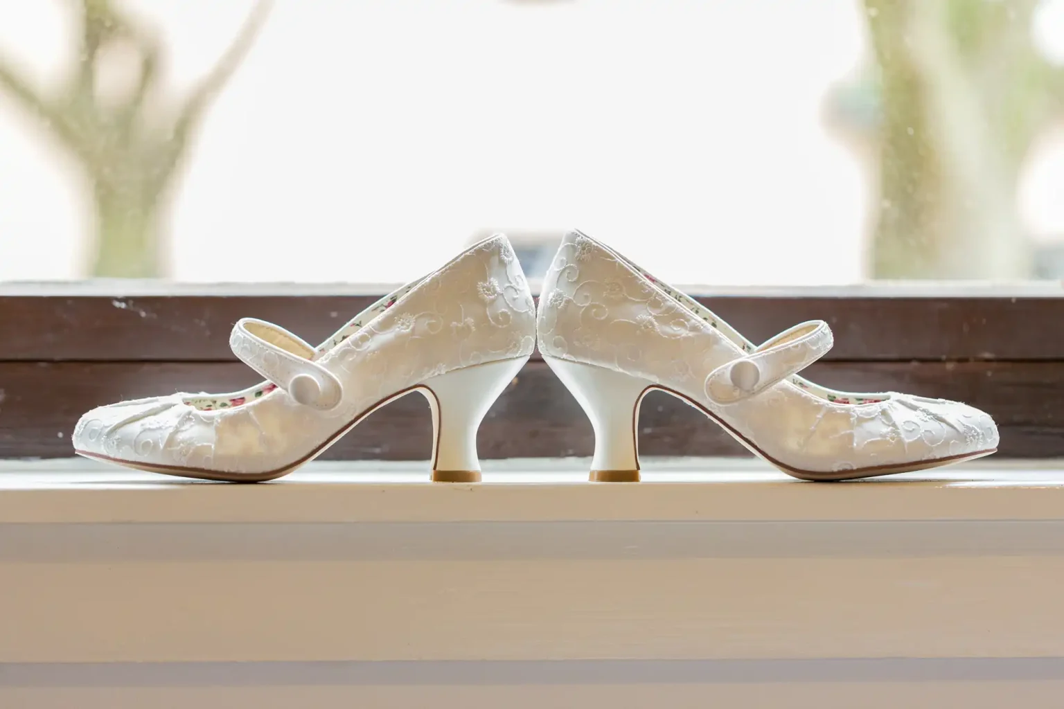 A pair of white, lace-embellished bridal shoes with medium heels, displayed on a windowsill against a blurred background.