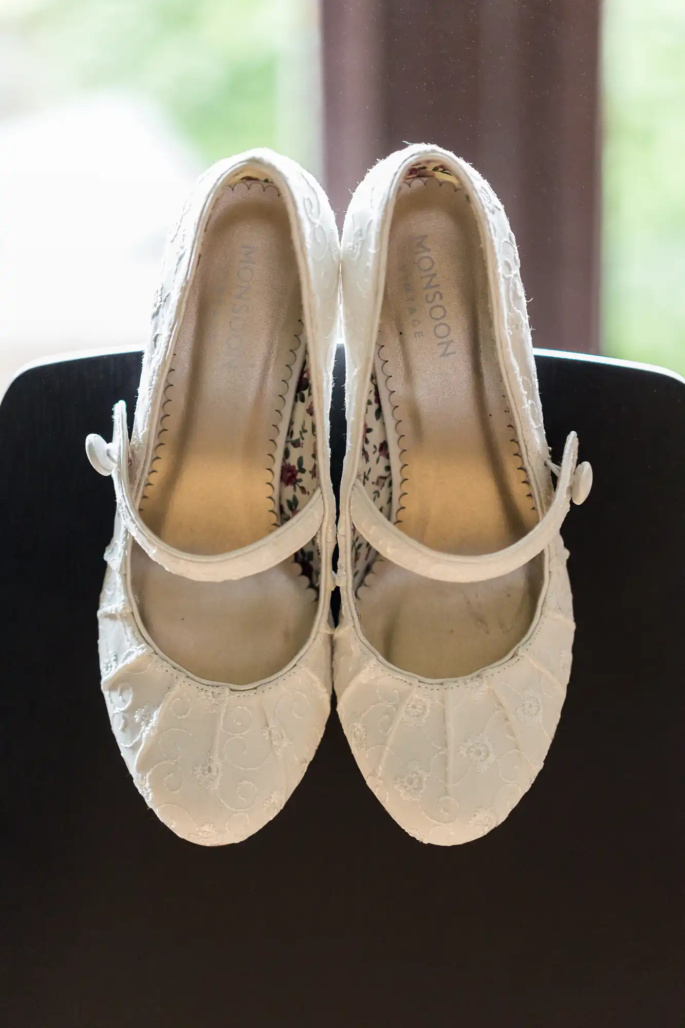 A pair of white, lace monsoon ballet flats positioned on a dark surface with a natural light background.