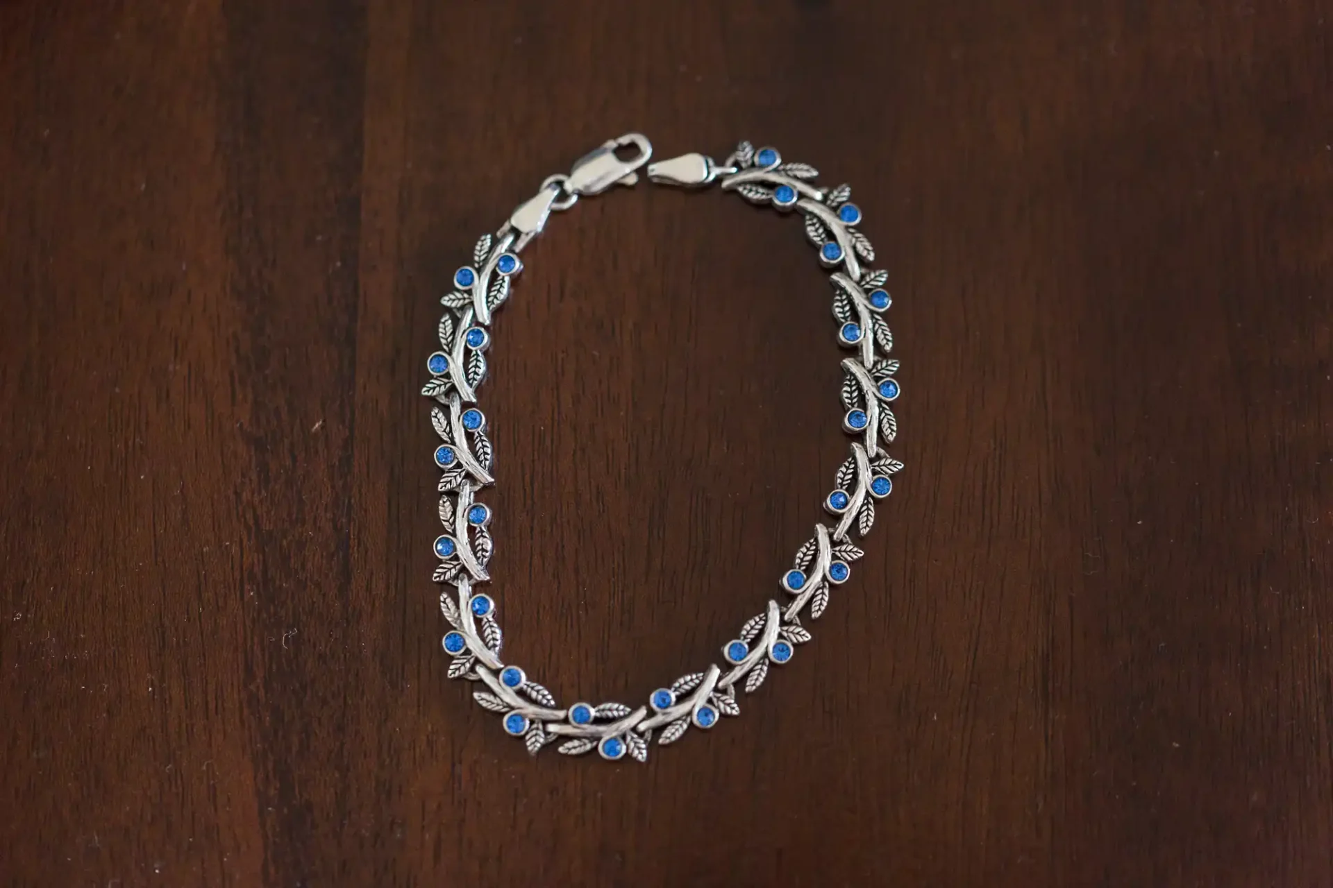A silver bracelet with blue and white gemstones, laid in a circular shape on a dark wooden surface.