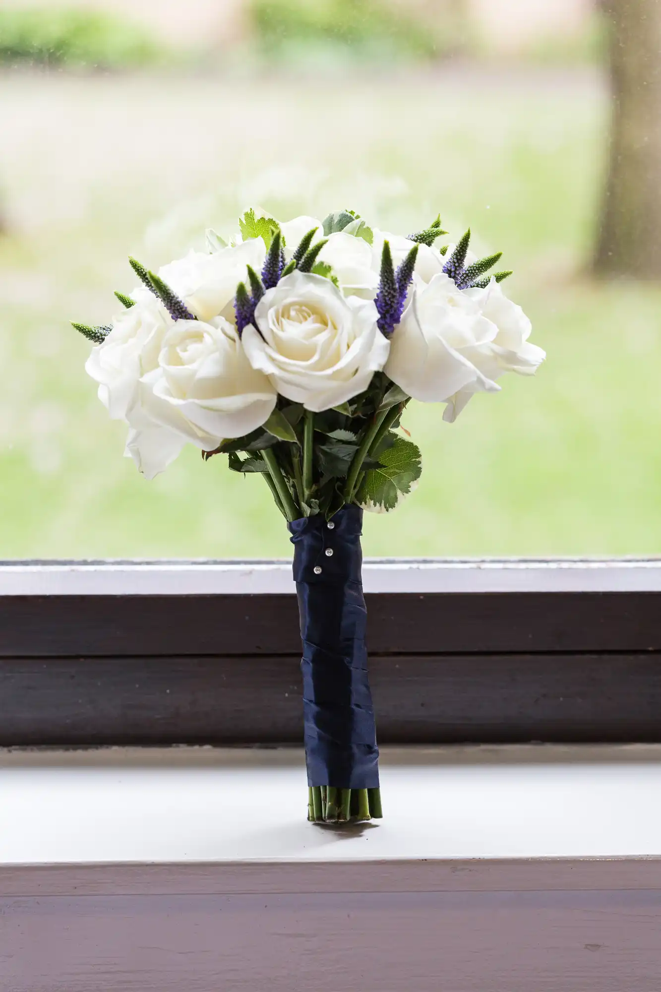 A bouquet of white roses and purple flowers tied with a navy ribbon, standing on a windowsill with a blurred green background.