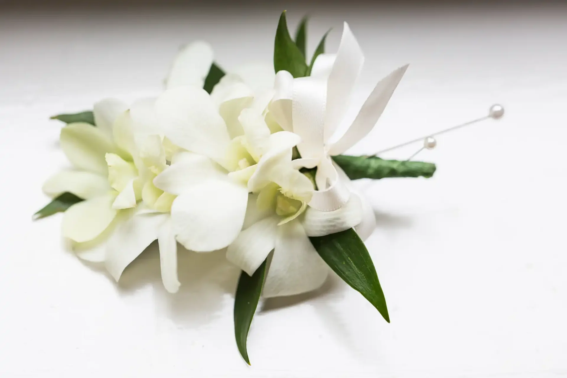 A delicate white floral corsage with green leaves and pearl accents on a white background.