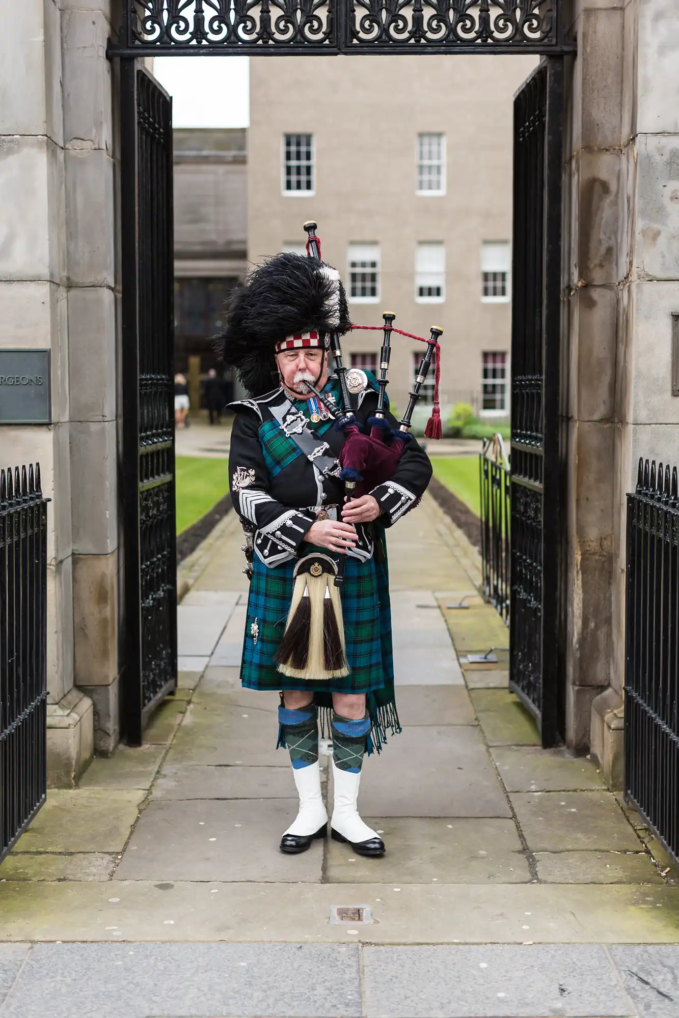 A bagpiper in traditional scottish attire, including a kilt and sporran, stands playing bagpipes at a gate entrance.