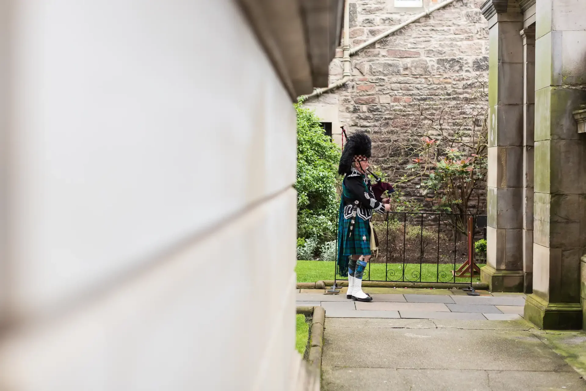 A bagpiper in traditional scottish attire playing in a historic courtyard, framed by blurred architectural details foreground.