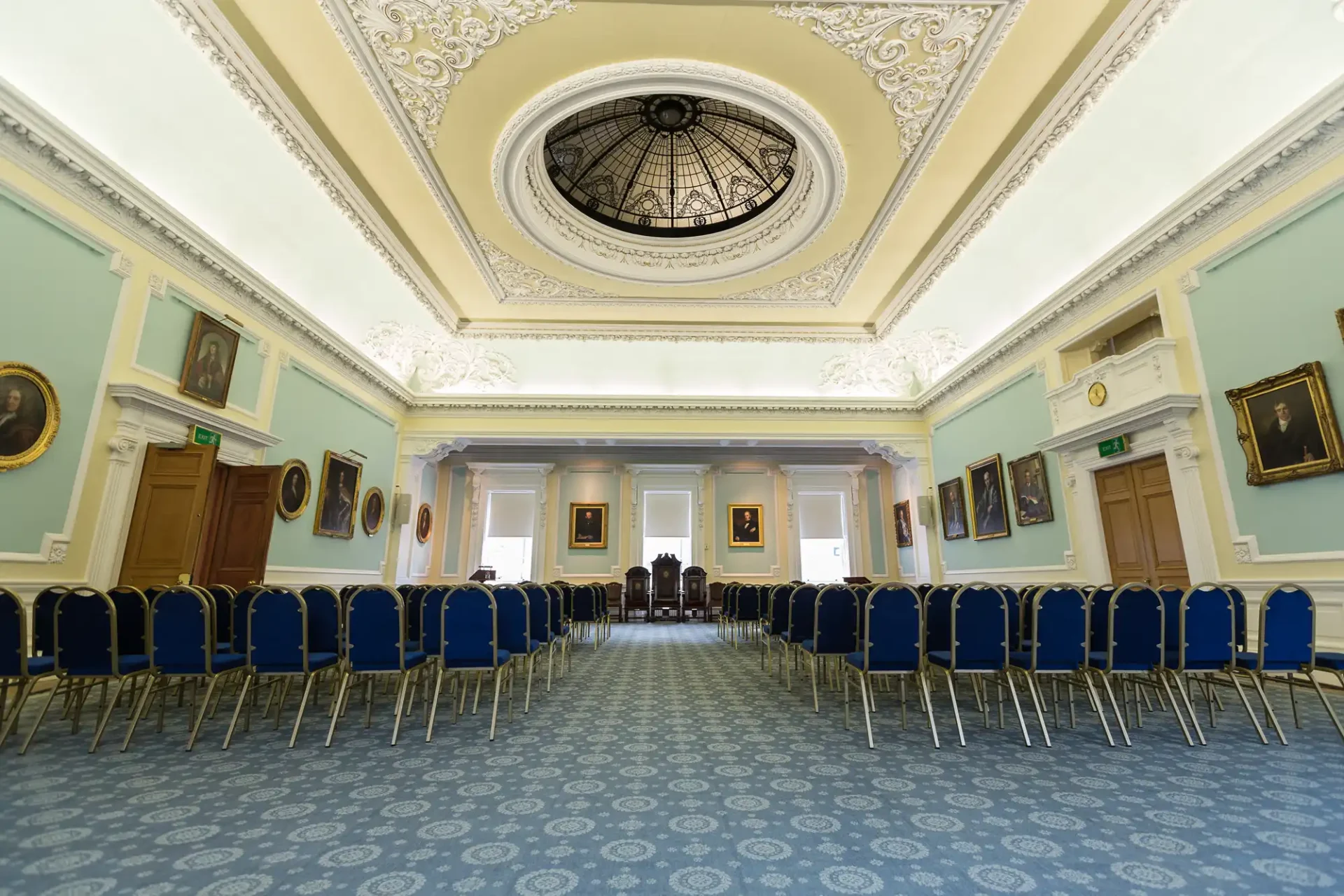 Elegant room featuring rows of blue chairs, light blue walls adorned with portraits, intricate white ceiling with a central skylight, and a patterned blue carpet.