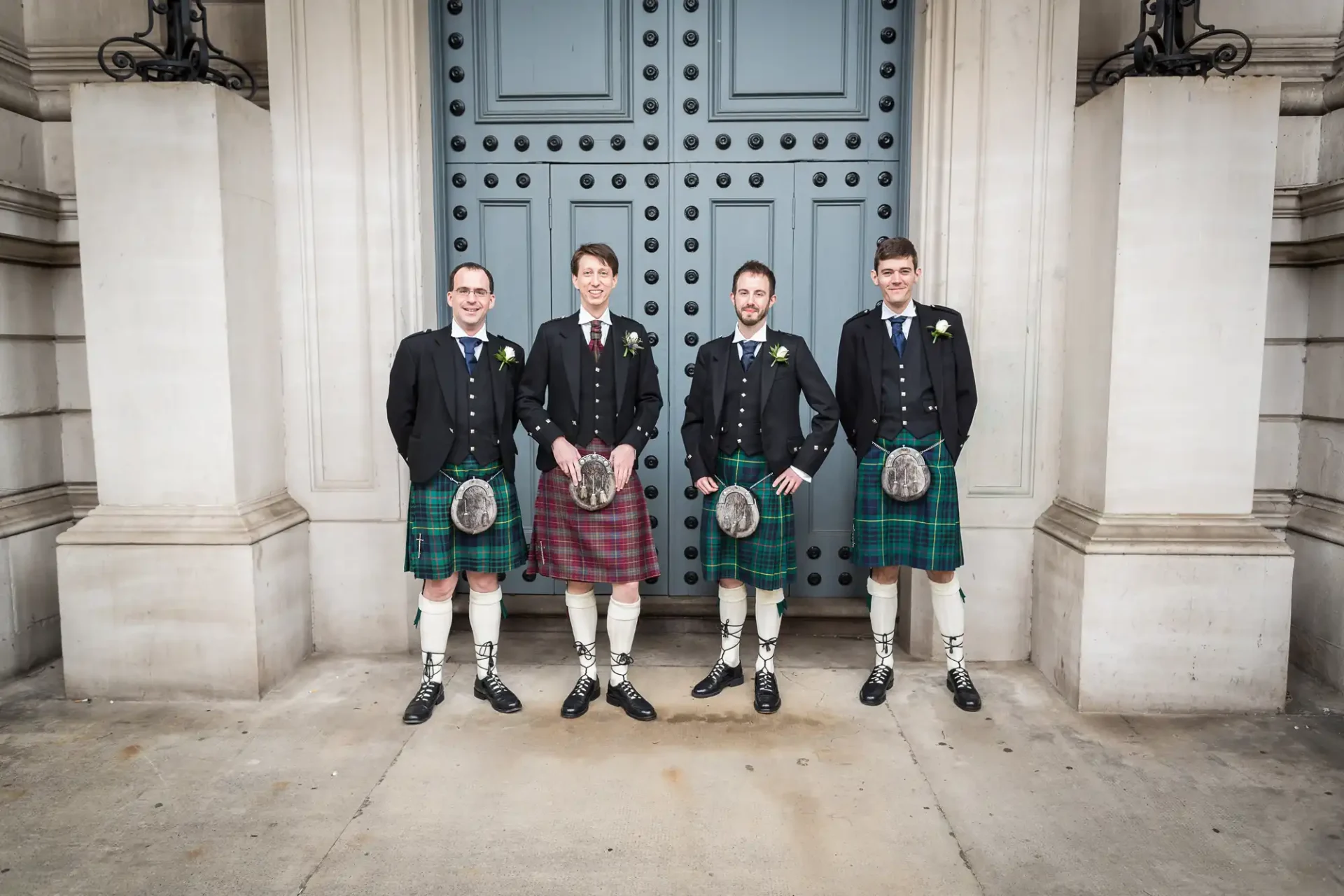 Four men in traditional scottish attire with kilts standing in front of an ornate building with blue doors.