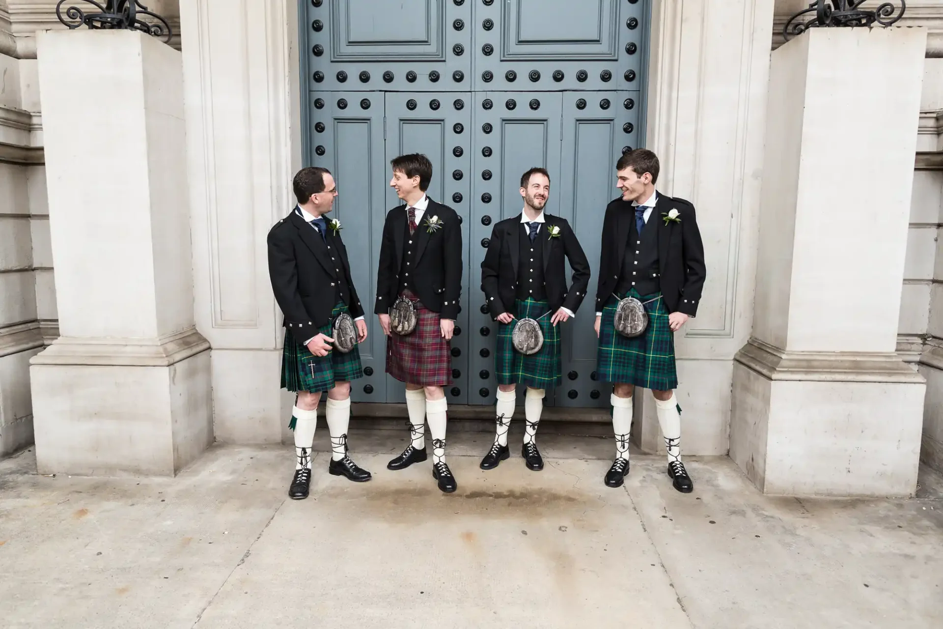 Four men in traditional scottish kilts and jackets, laughing and talking in front of a large blue door.