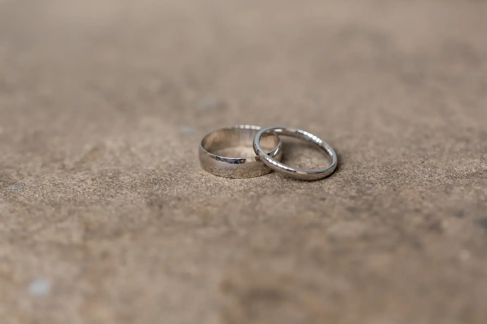 Two wedding rings lie on a textured surface, one slightly in front of the other, with a soft-focus background.