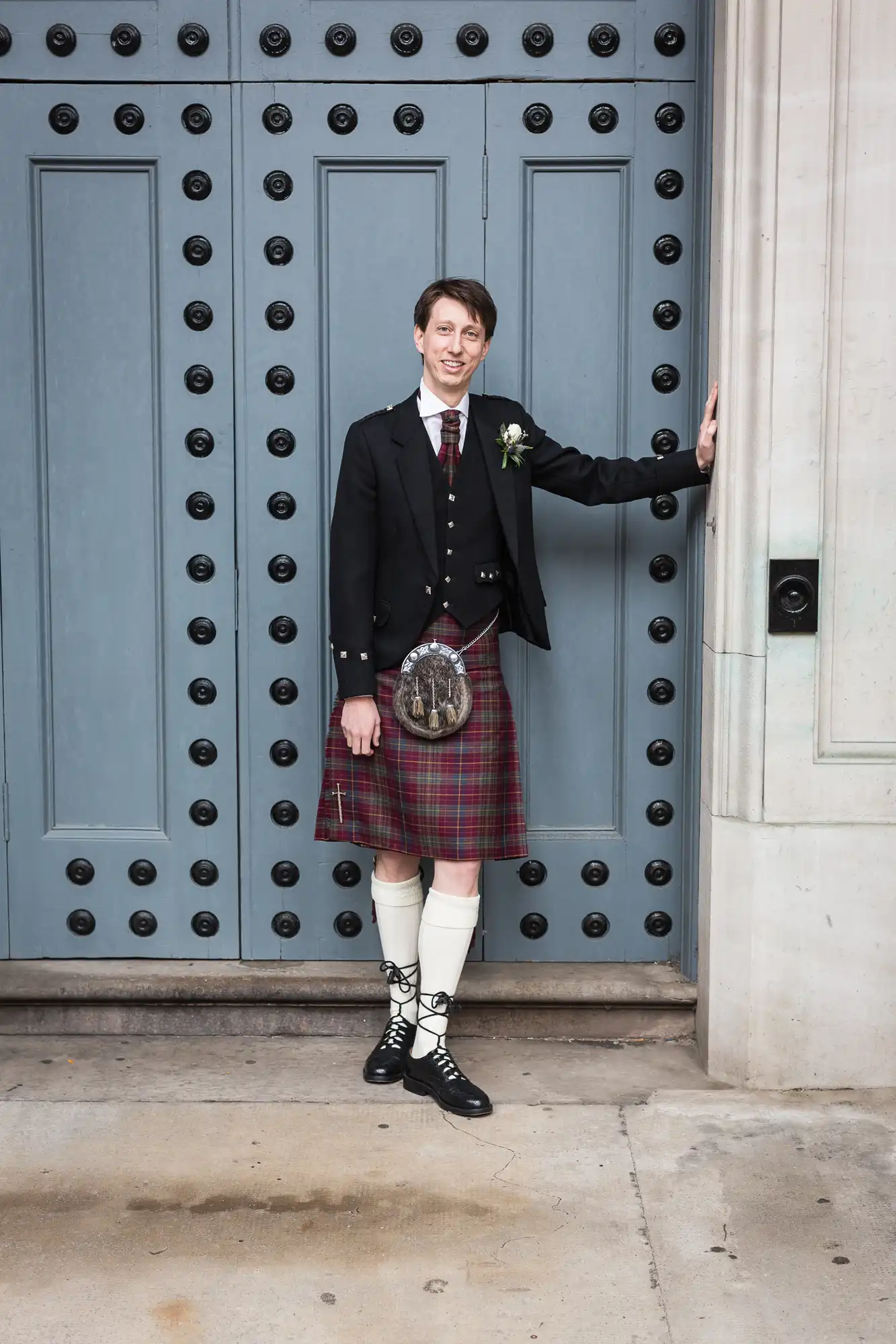 A man in traditional scottish attire, including a kilt and sporran, stands by a large blue door, smiling and holding the door handle.