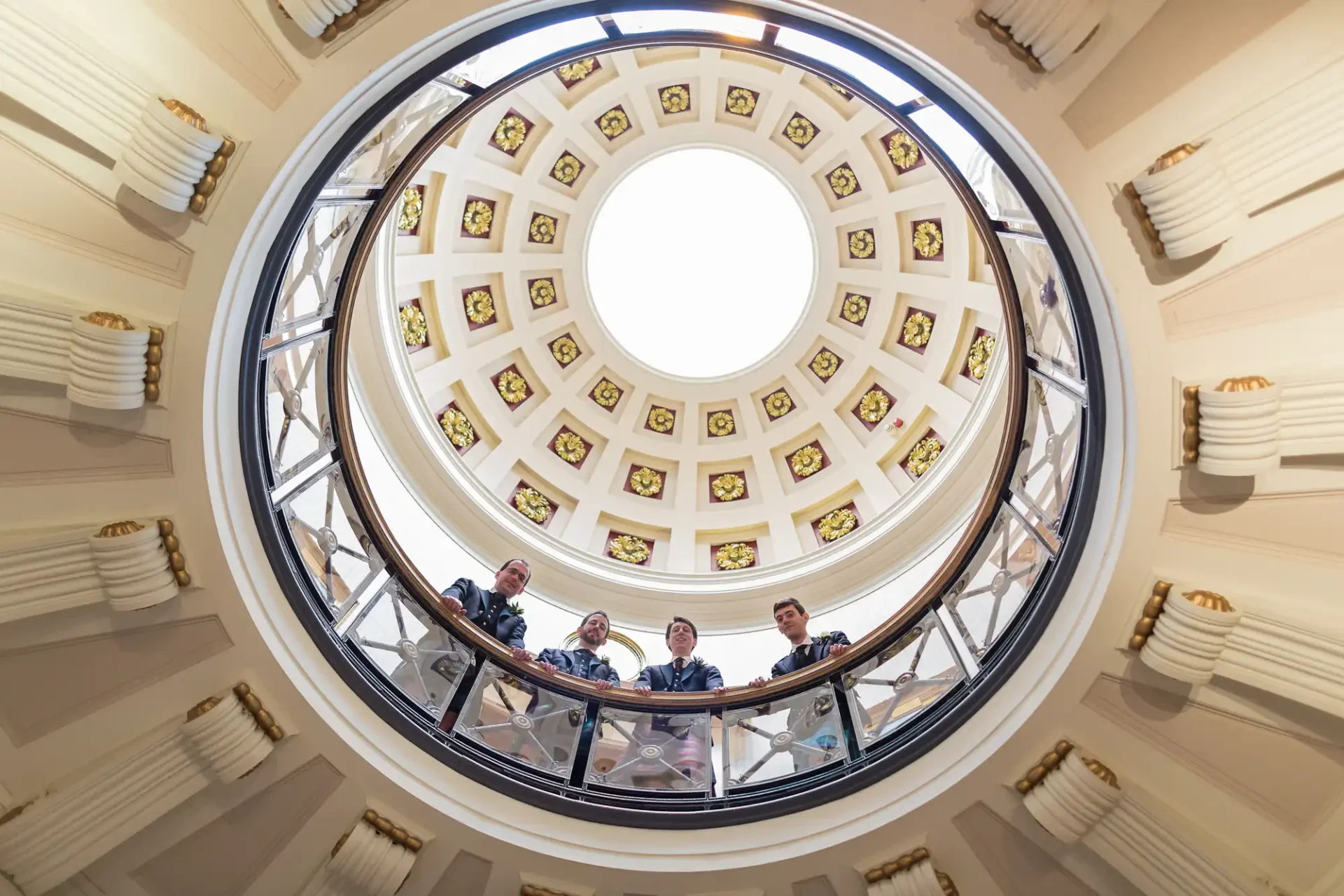 Four people standing on a balcony under a large circular skylight in an ornate building with neoclassical architecture.