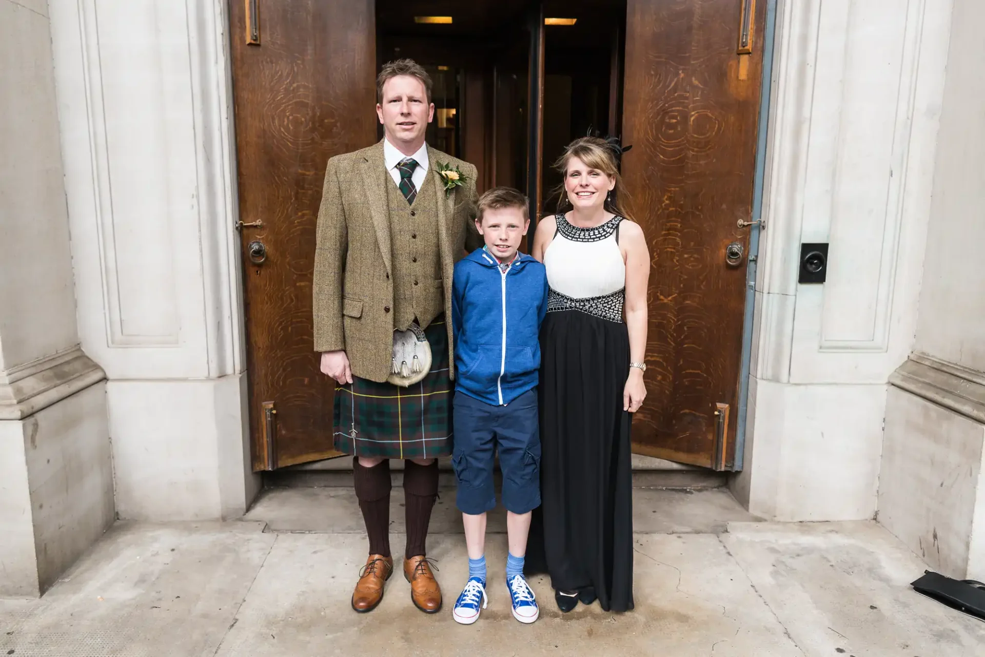 A family of three standing in front of a building entrance; the man wears a kilt, the boy is in a suit with sneakers, and the woman is in a long black dress.