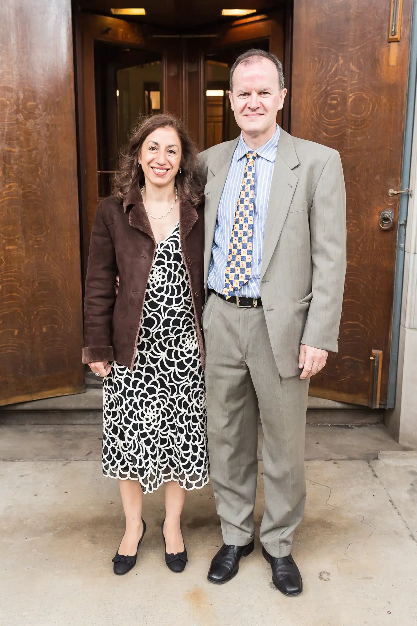 A man and a woman standing and smiling in front of wooden doors, dressed in formal attire, the man in a suit and the woman in a dress and jacket.