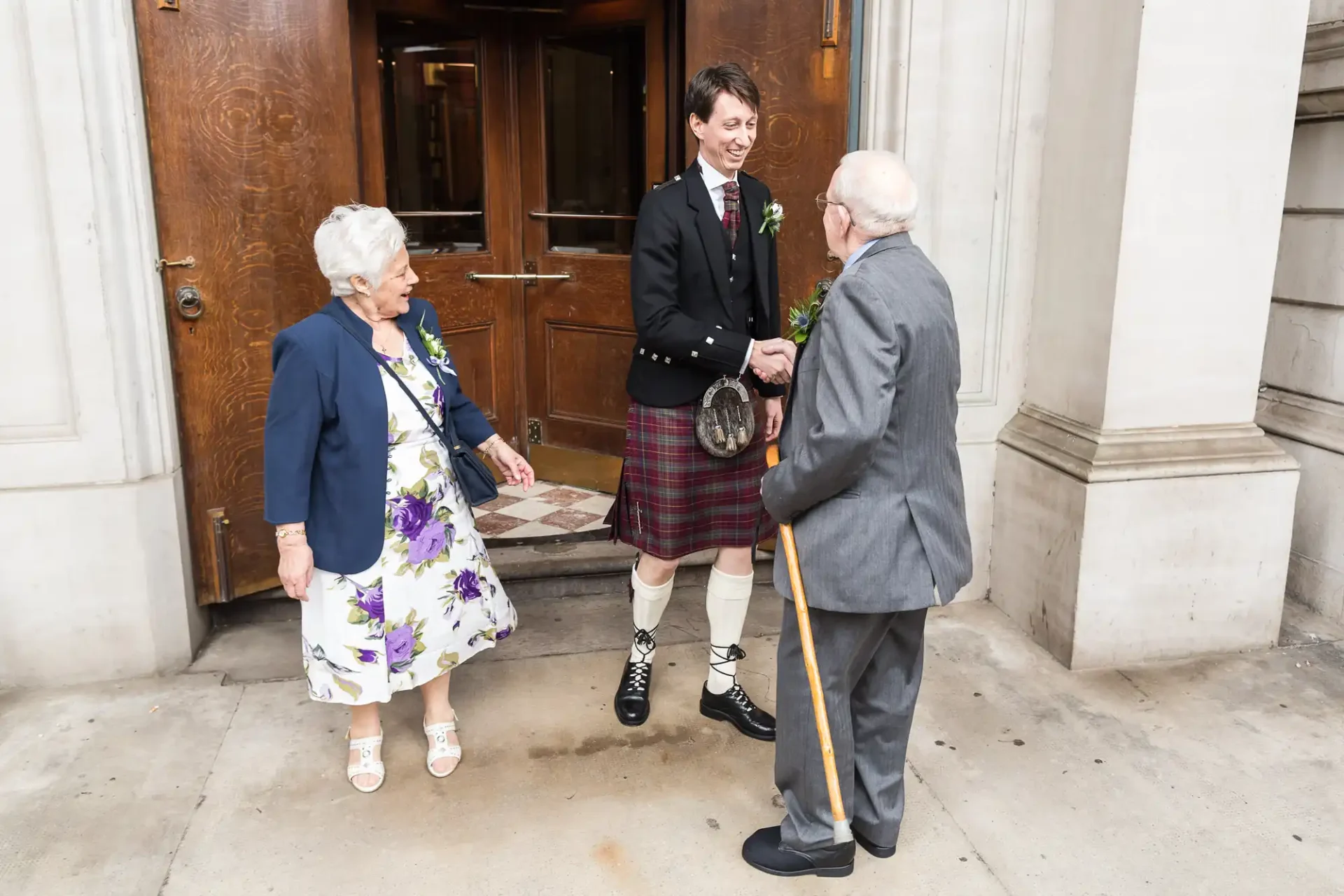 A groom in traditional scottish attire greets an elderly man with a cane outside a building, with an older woman beside them looking on.