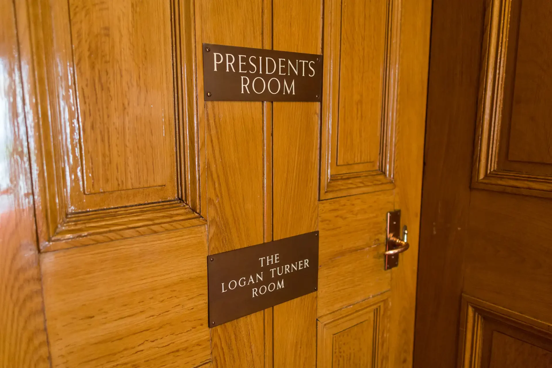 A wooden door with two signs reading "presidents room" and "the logan turner room," featuring a stainless steel handle.
