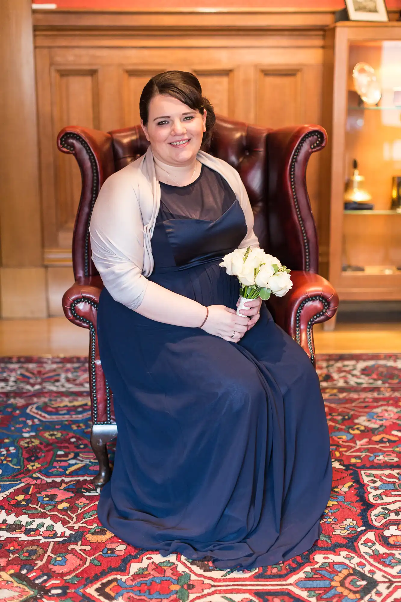 A woman in a navy blue evening gown and white shawl, holding a bouquet of white roses, seated in a red and blue patterned armchair.
