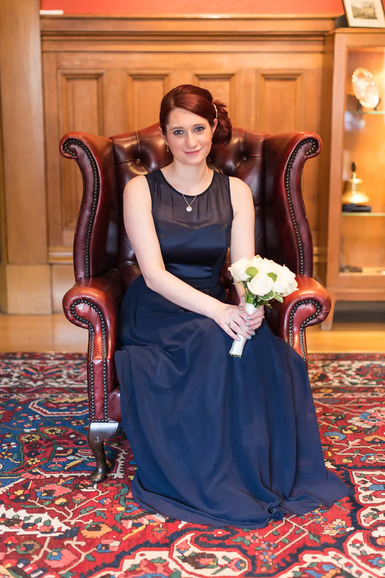A woman in a navy blue gown sits on a vintage chair, holding a bouquet of white roses, with a patterned carpet beneath her.