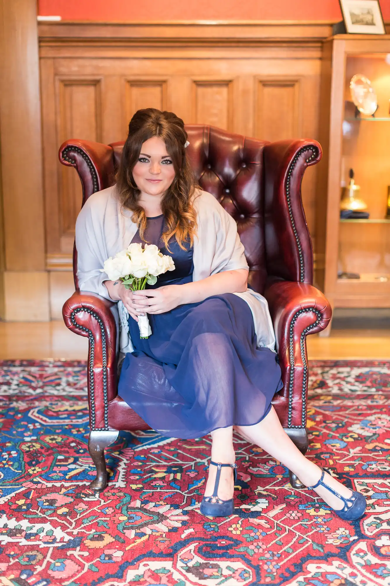A woman in a navy blue dress and matching shoes sits in a red leather armchair, holding a bouquet of white flowers, with a patterned red and blue rug underneath.