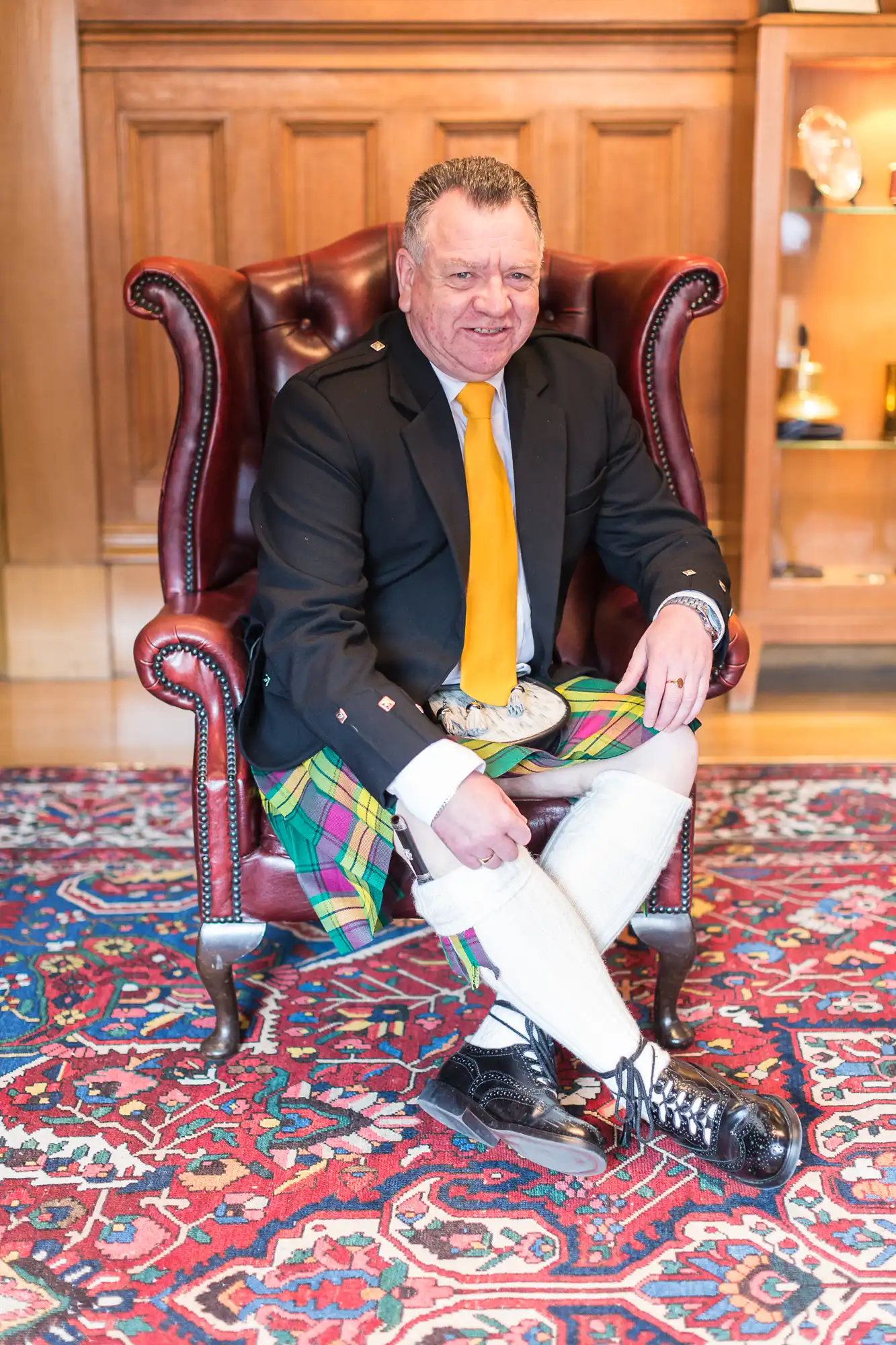A man in a traditional scottish outfit, including a kilt and sporran, sits confidently in a leather chair against a richly patterned rug.