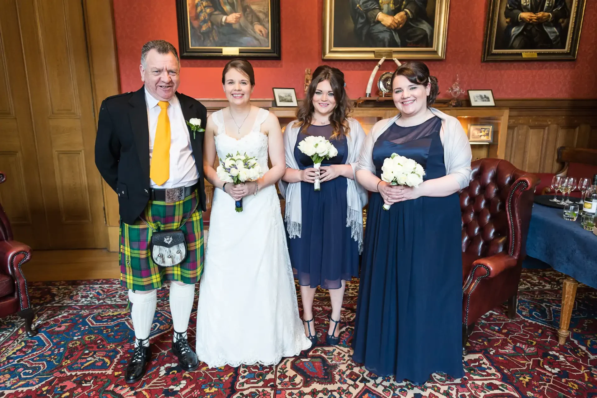 A bride and groom flanked by two bridesmaids holding bouquets in a room with dark wood paneling and paintings. the groom wears a traditional kilt.
