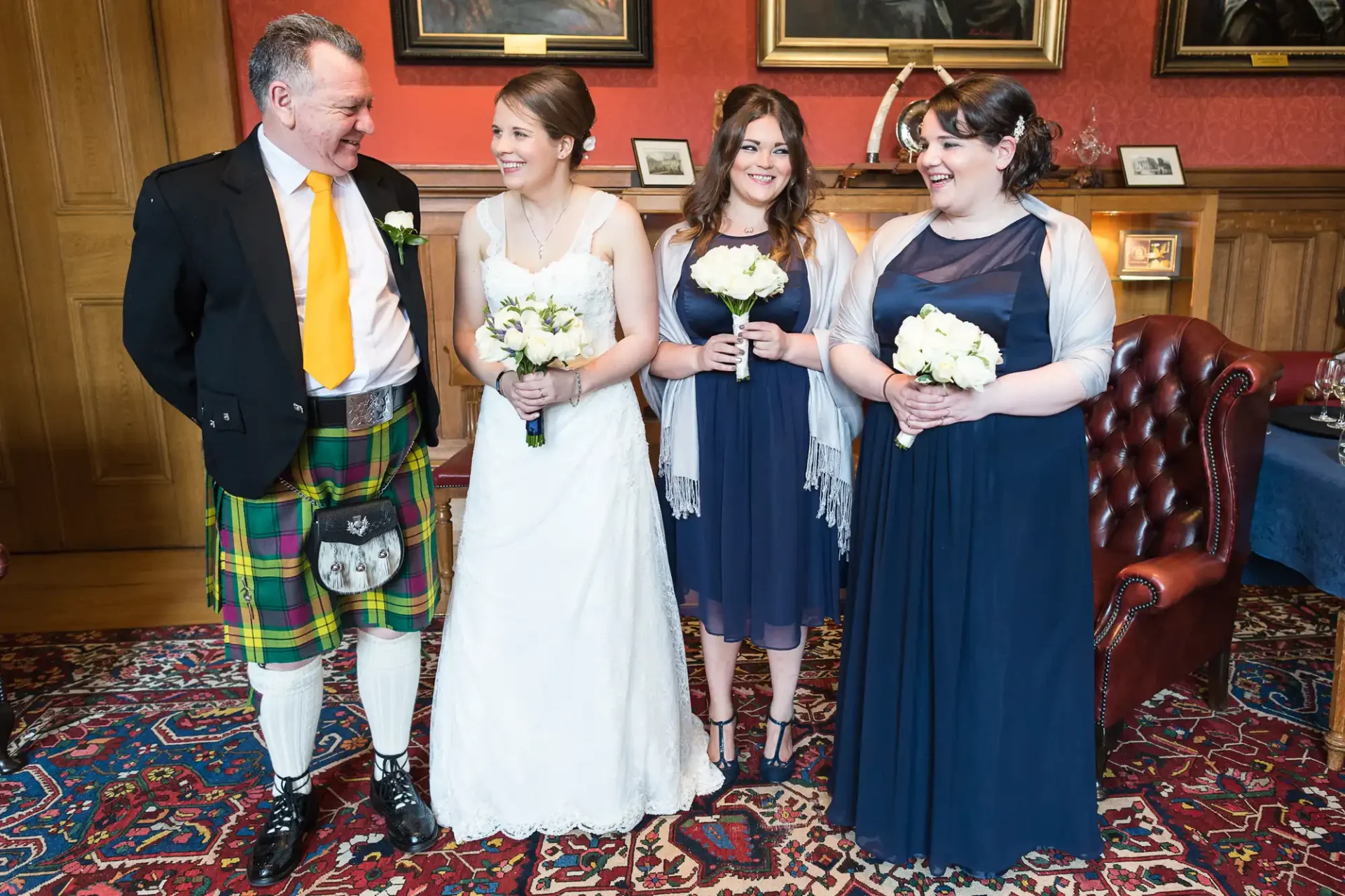 A bride in a white dress stands with her father in a tartan kilt and two bridesmaids in navy dresses, all holding bouquets, in an elegant room.