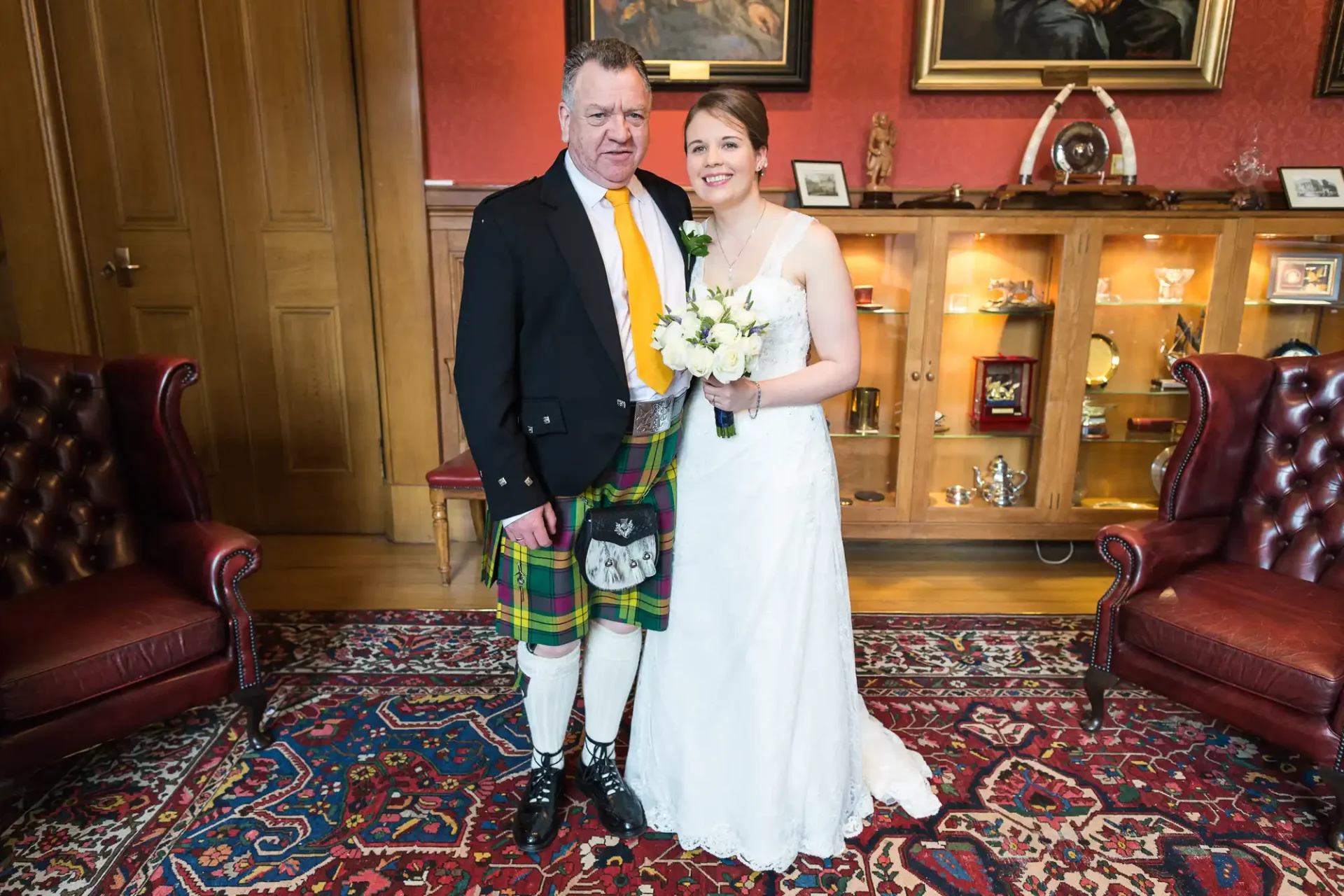 A bride and groom pose for a photo indoors; the groom wears a tartan kilt and the bride is in a white gown, holding a bouquet. they stand smiling between two red leather chairs on a patterned carpet.