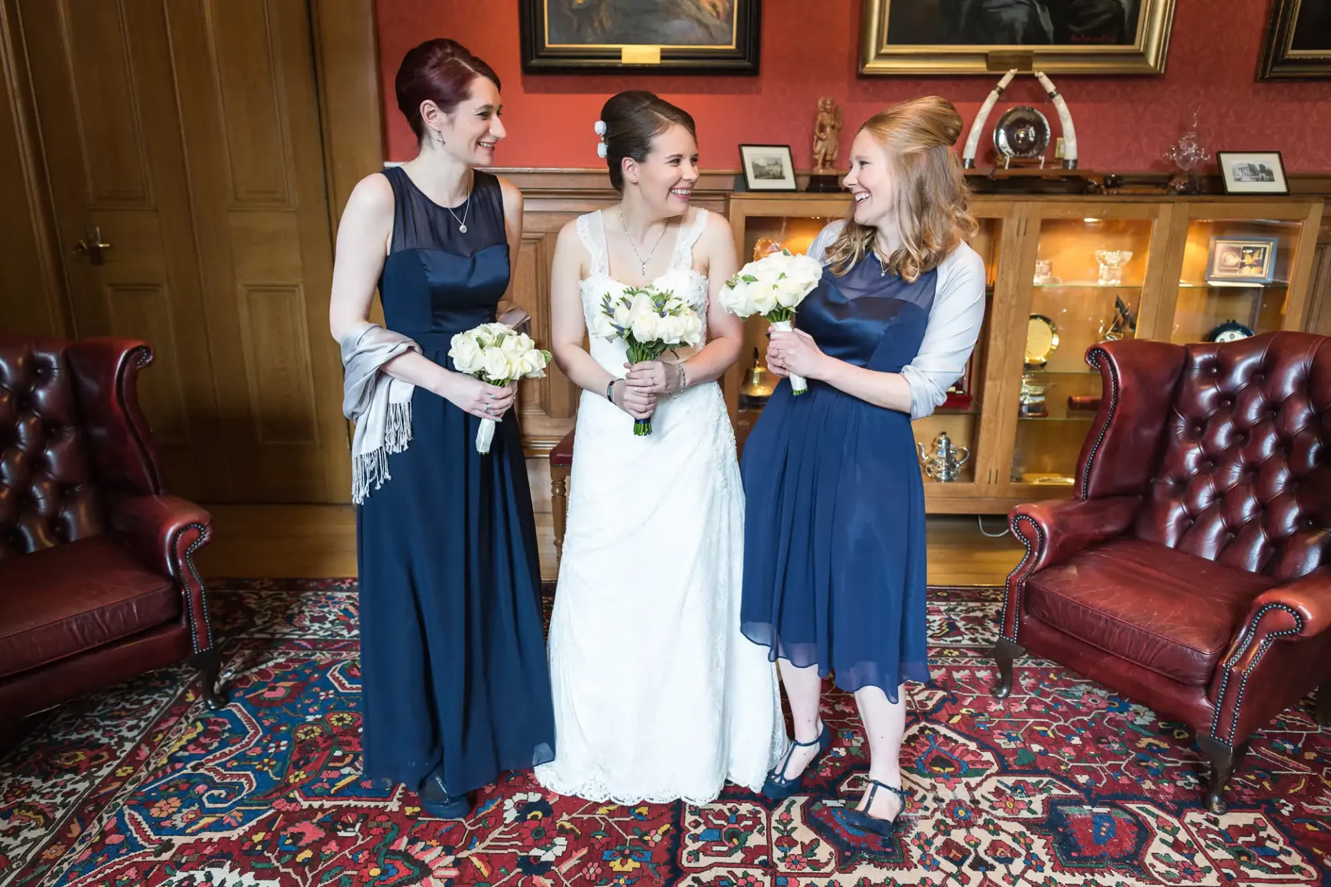 Three women in formal attire, two in navy dresses and one in a white wedding dress, holding bouquets and smiling in an elegant room with red chairs and a patterned rug.