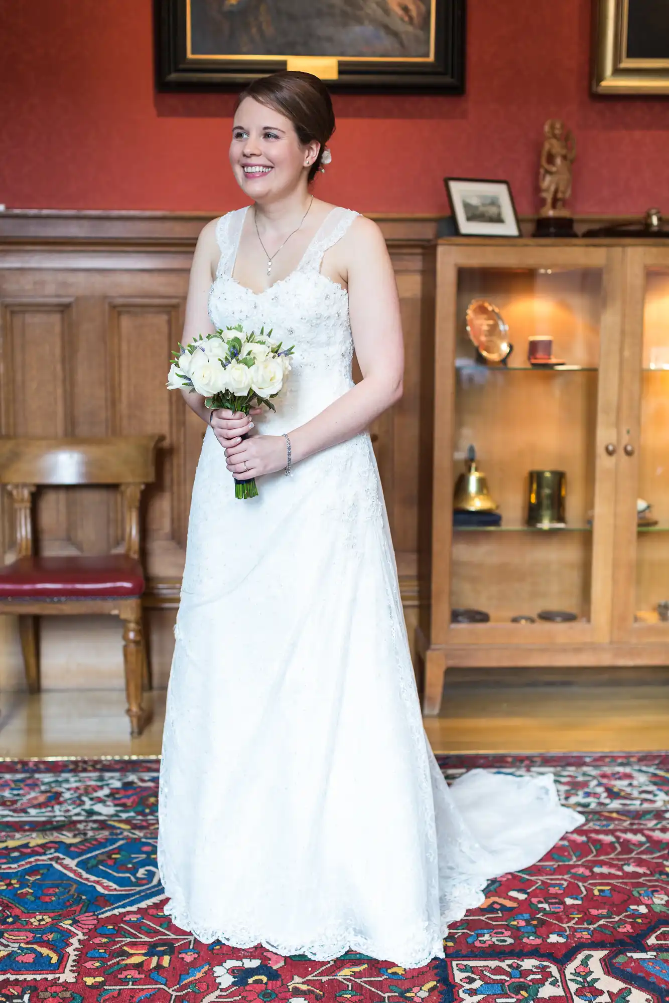 A bride in a white lace gown holding a bouquet of white flowers, standing in a room with a wooden cabinet and a red patterned carpet.