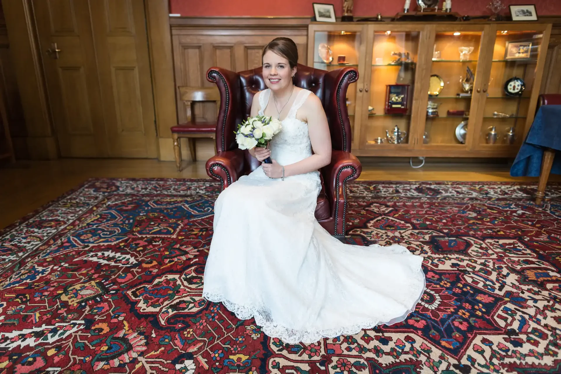 A bride in a white dress smiles while sitting in a red leather chair, holding a bouquet, with a cabinet and persian rug in the background.