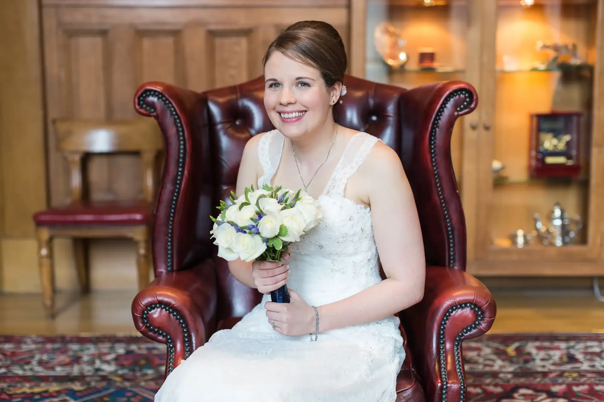 A bride in a white dress sits smiling on a red leather chair, holding a bouquet of white and pale yellow flowers, with a wood-paneled background.